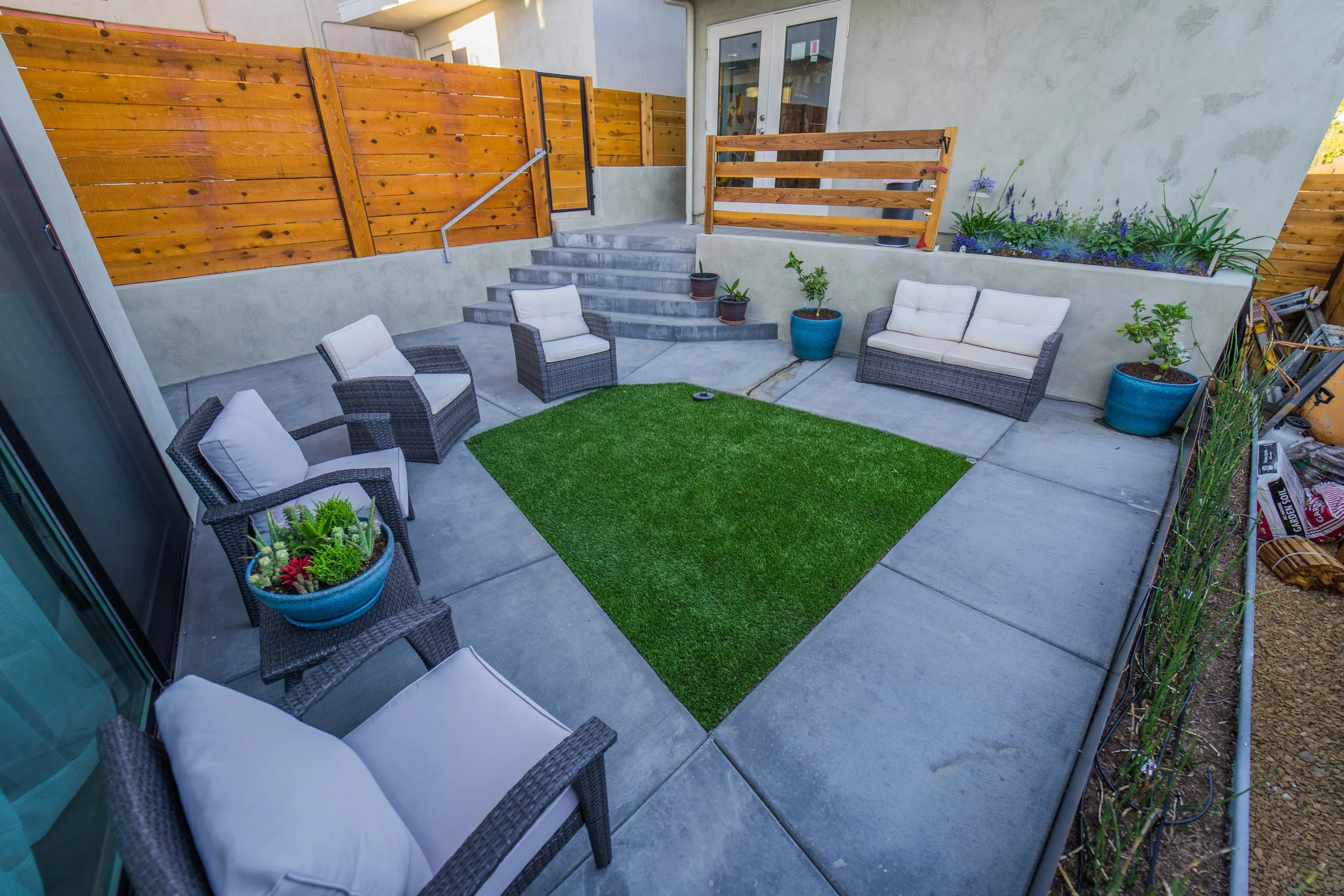 Small outdoor patio area with gray concrete flooring, a grassy patch, and patio furniture including a white cushioned loveseat and two armchairs with gray wicker frames. There are potted plants including succulents and small trees, a wooden staircase with a metal handrail leading up to a doorway, and a raised flower bed with purple and green plants. Wooden fencing surrounds part of the patio, and some gardening tools are visible on the right side.