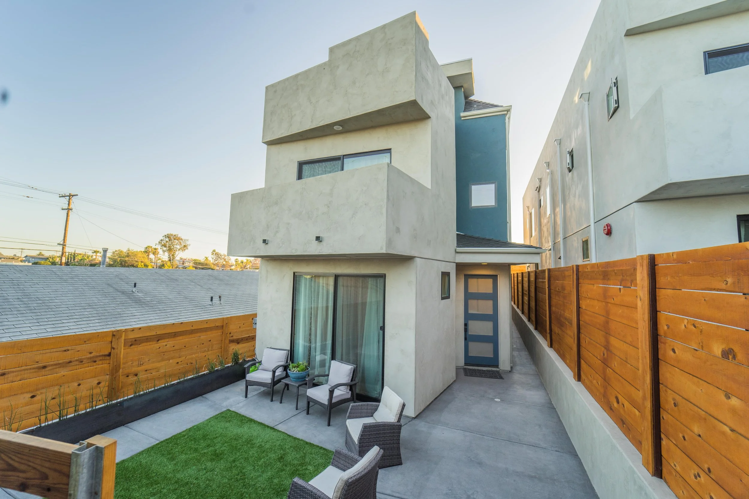 Small urban backyard patio with patio chairs, a sliding glass door, a wooden fence, and a modern multi-story apartment building in the background.