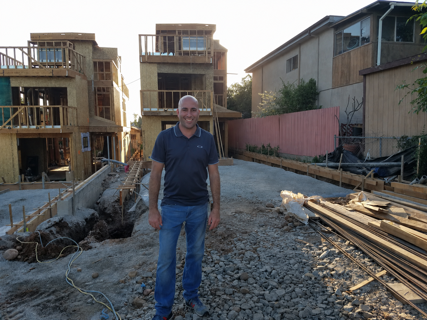 Man standing on a construction site with new wooden-framed buildings in the background, construction materials and debris on the ground.