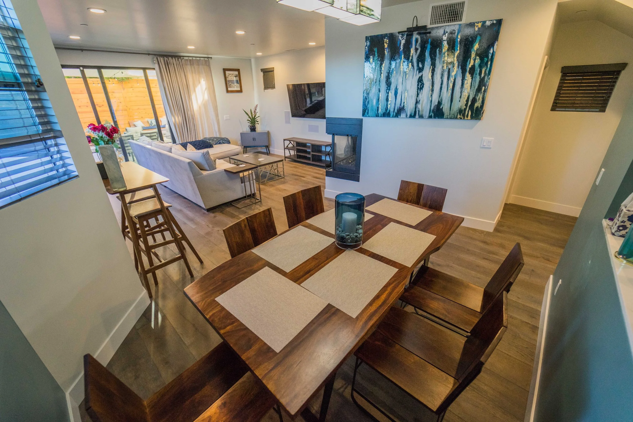 View of a modern living room and dining area with wooden furniture, a floral arrangement, and wall art, with large sliding glass doors leading to an outdoor patio.