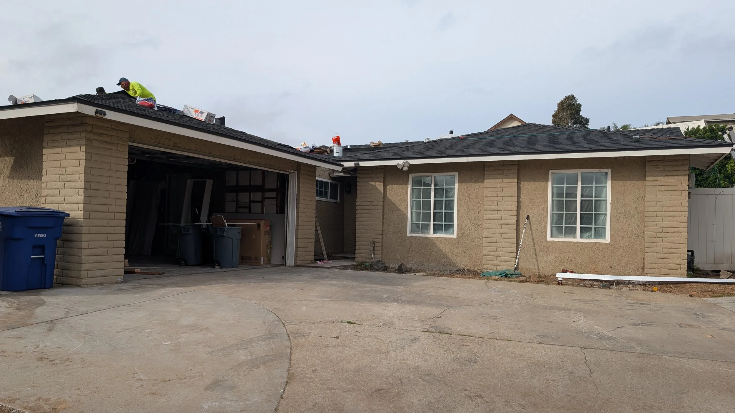 A house with a person working on the roof, visible roofing materials and tools on the roof, two large windows, a garage with open door, trash bins, and a driveway, during daytime with a cloudy sky.