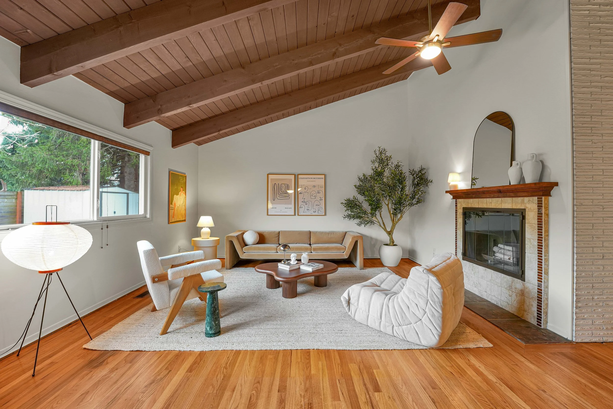 Living room with wood-paneled vaulted ceiling, ceiling fan, large window, standing lamp, several framed art pieces on the wall, a potted plant, fireplace, and modern furniture including a beige couch, white armchair, and a wooden coffee table.