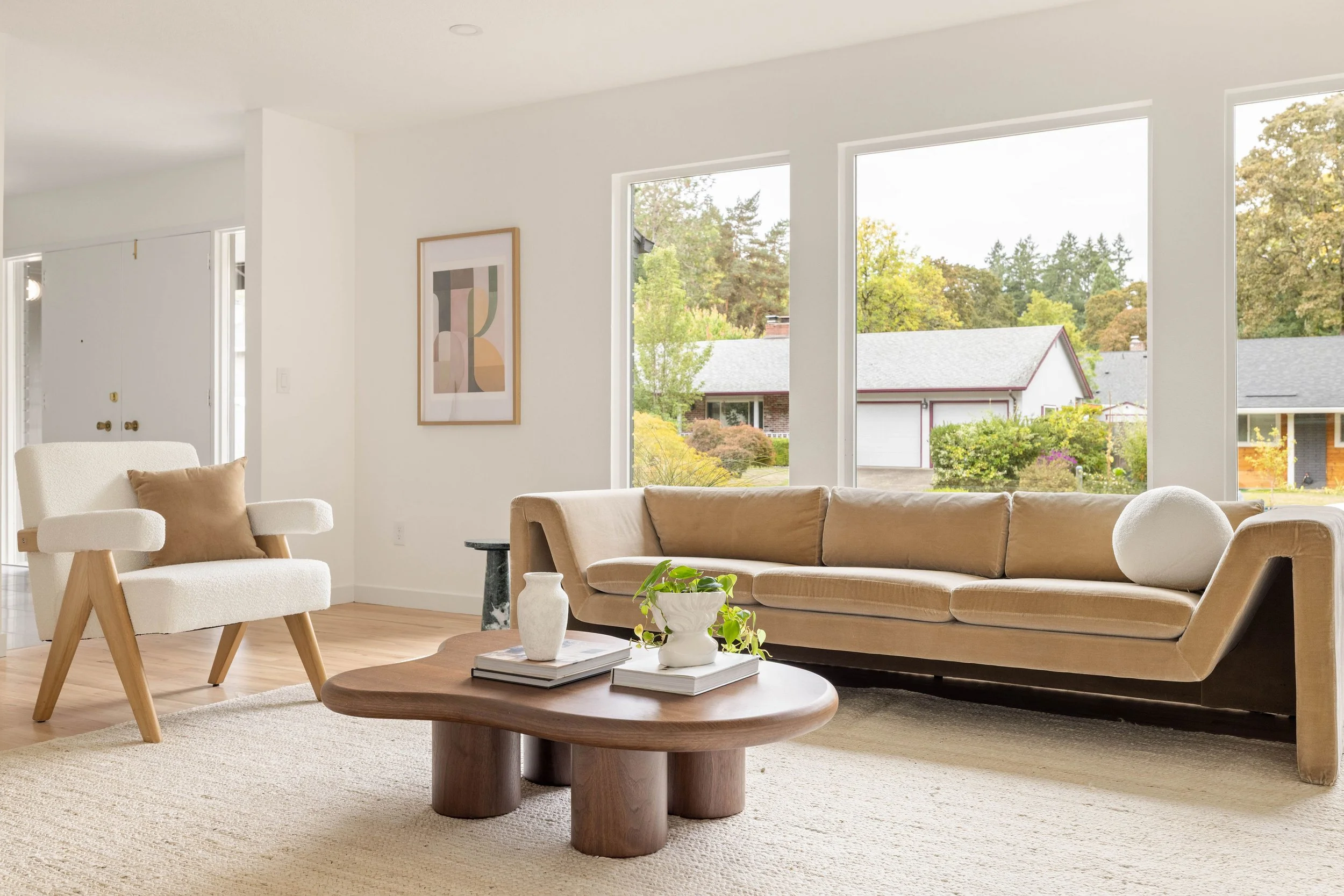 Living room with white walls and large windows, beige sofa, white armchair with cushion, wooden coffee table with vases and books, abstract wall art, viewed outdoors of neighborhood with trees and houses.