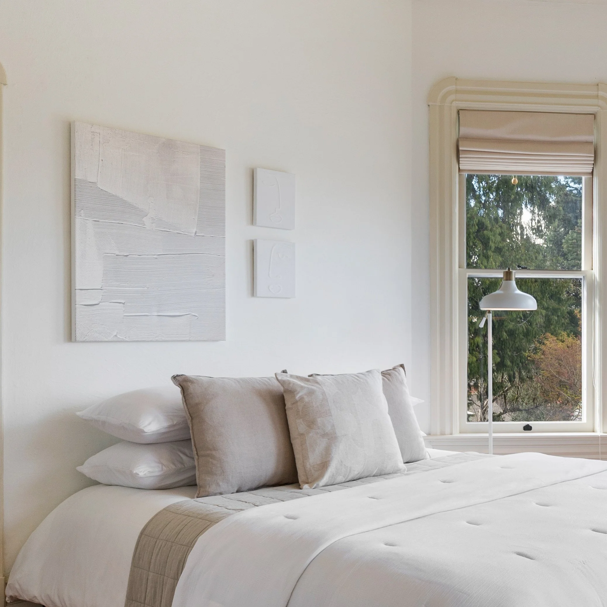 Neatly made bed with white bedding and beige pillows in a bright bedroom with large window overlooking trees and sky.