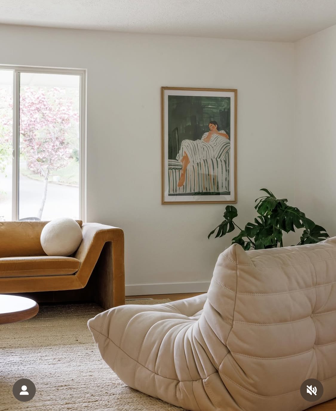 Living room with beige couch, white armchair, green potted plant, framed artwork of a woman on a striped sofa, and window showing trees with pink blossoms.