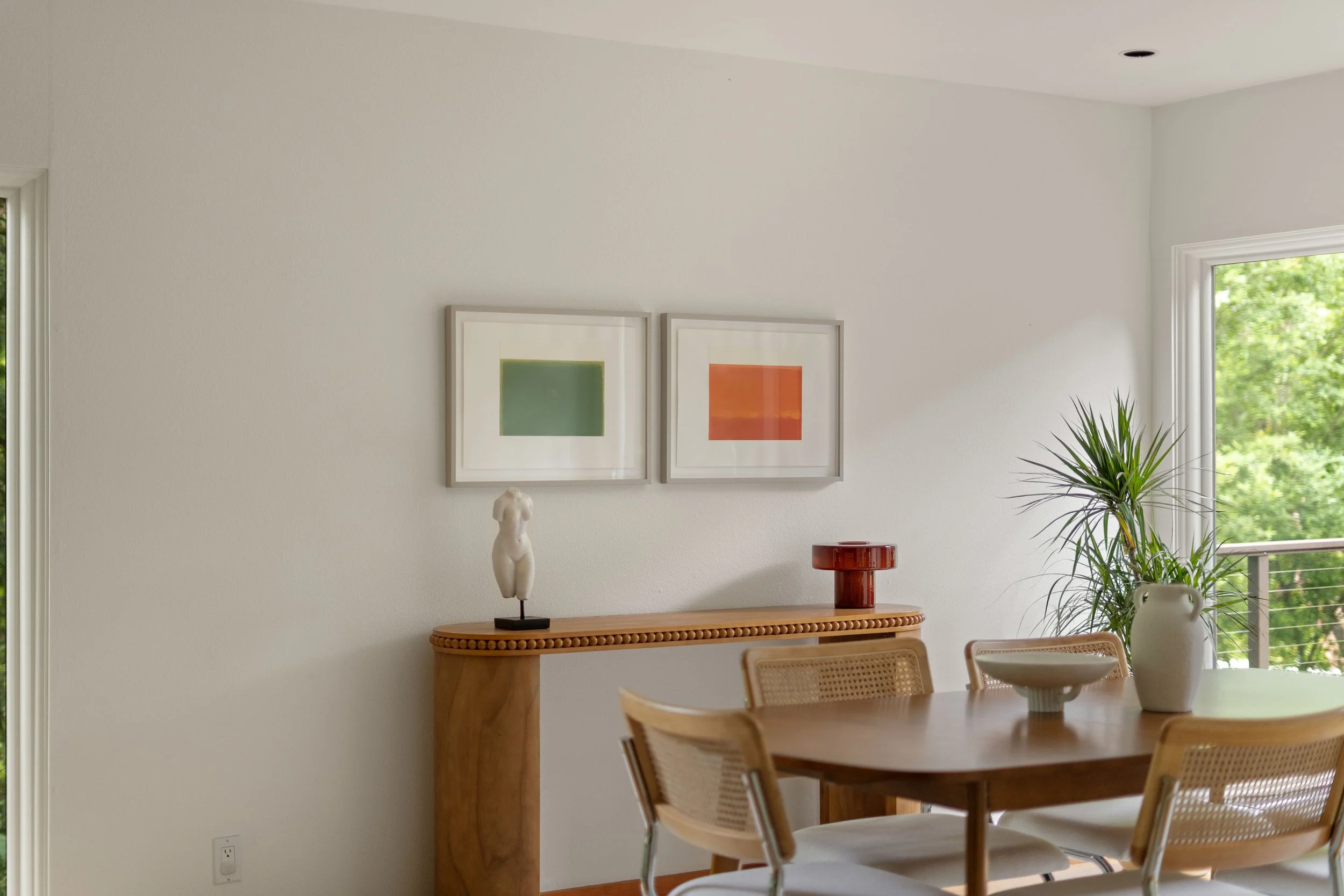 A dining room with a wooden table and beige chairs. On the wall behind, there are two framed abstract artworks, a sculpture on a console table, a potted plant, and a window showing green trees outside.