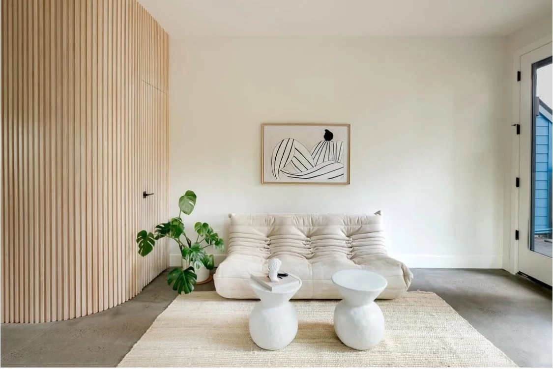Minimalist living room with a beige sofa, a potted plant, two white sculptural side tables, a wall-mounted abstract art piece, a wooden accent wall, and a sliding glass door.