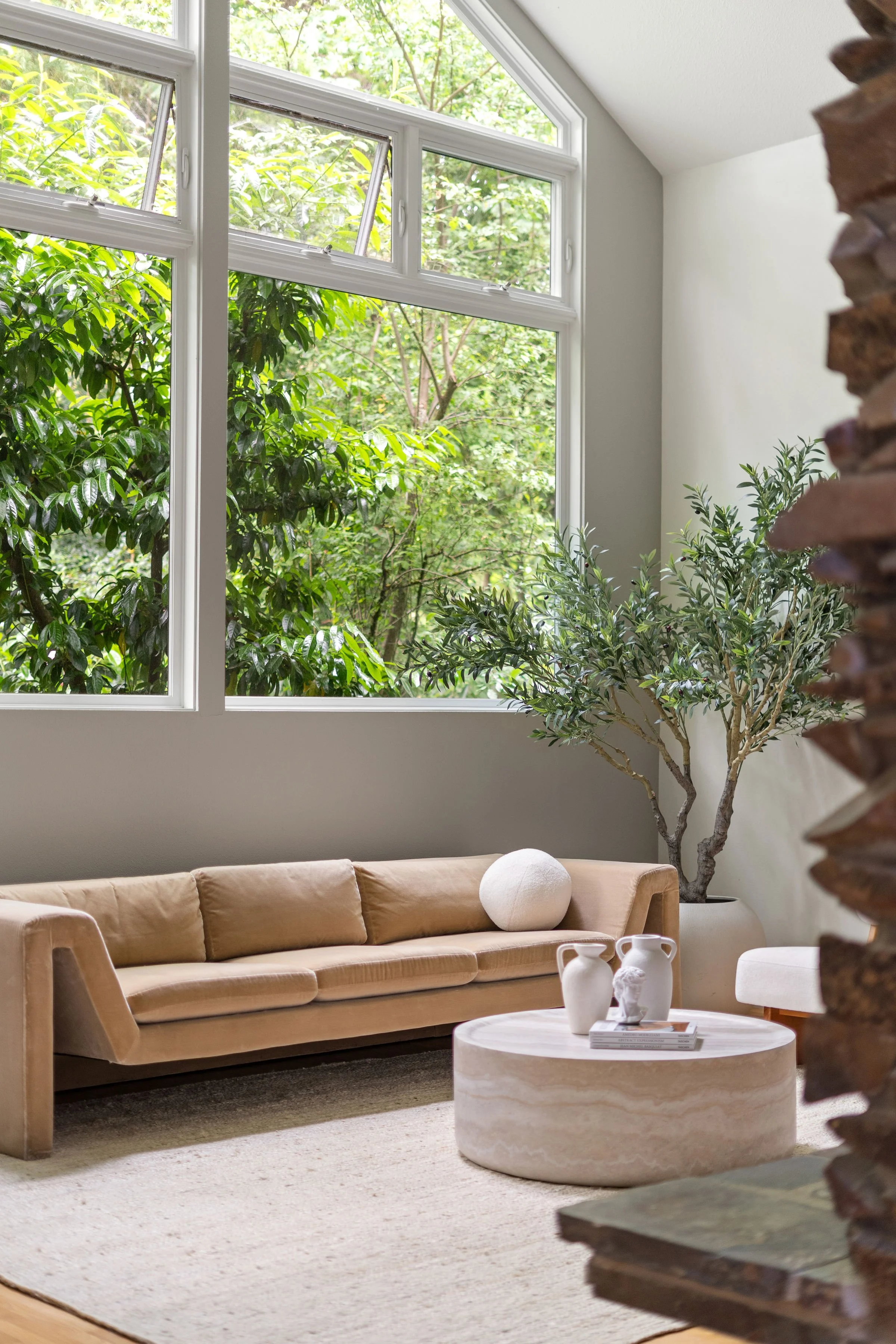 Living room with a beige sofa, white round coffee table with white vases, large window with green trees outside, indoor plants, and a brick wall on right