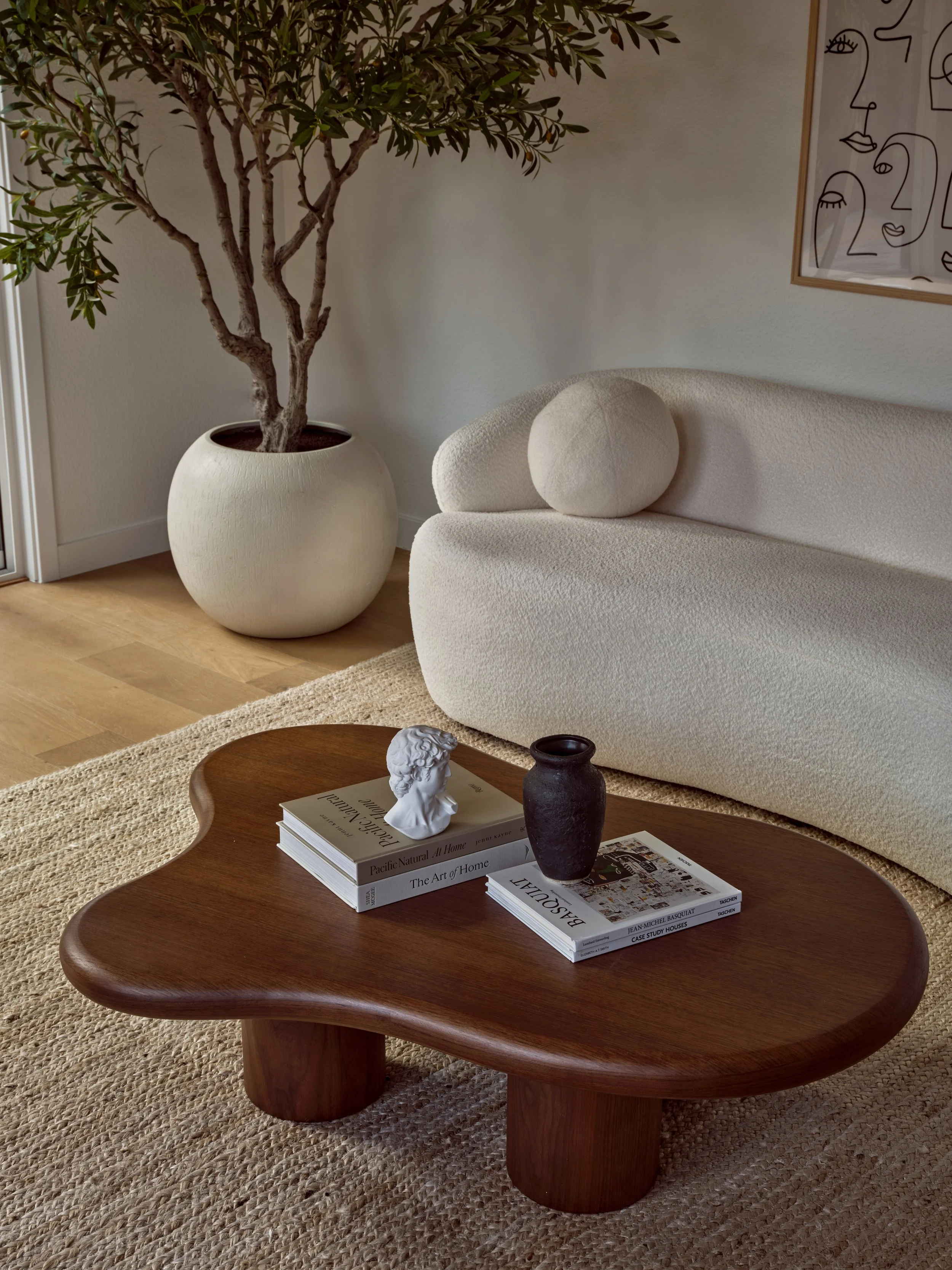 A living room with a large potted plant, a beige sofa with round cushions, a wooden coffee table with stacked books, and decorative items including a bust and a vase, with artwork on the wall.