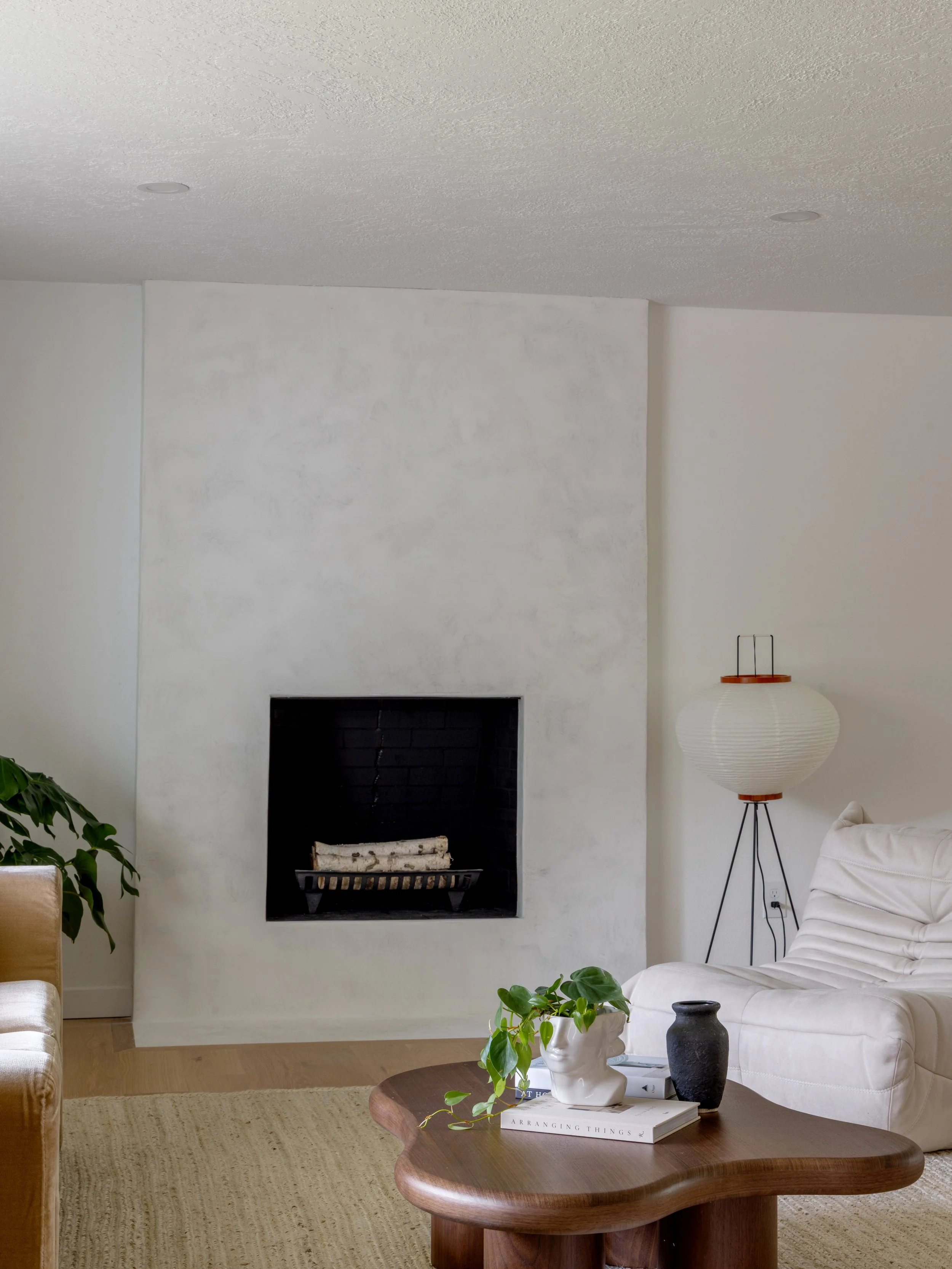 Living room with a fireplace, a floor lamp, a beige sofa, a coffee table with books and vases, and a textured wall.