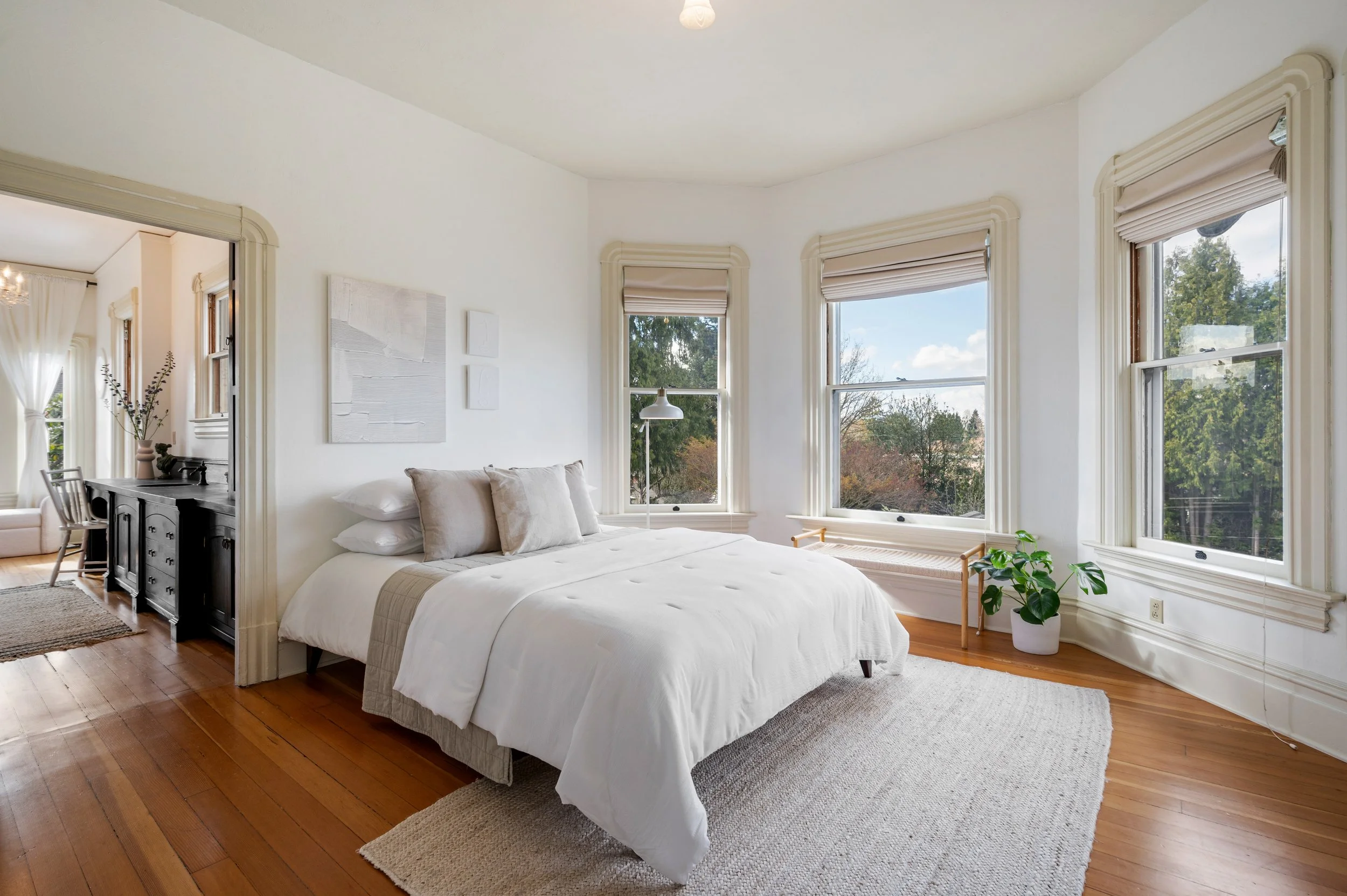 A bright bedroom with a white bed and beige pillows, surrounded by large windows showing trees and a blue sky, with a potted plant near the window and minimalist wall art.