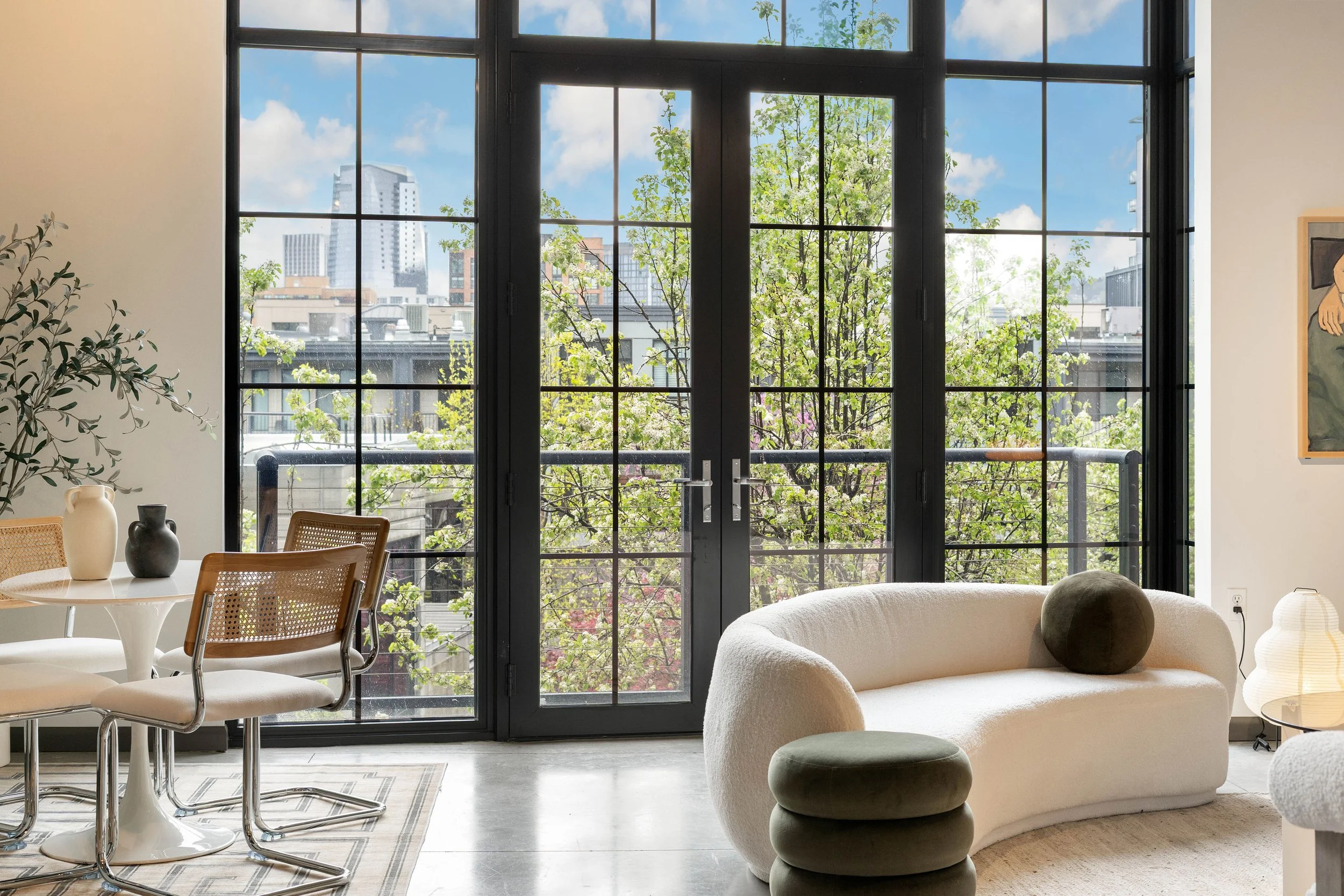 Living room with large black-framed windows showing trees and city buildings, modern sofa with pillows, round ottomans, a dining area with chairs, and decorative vases.