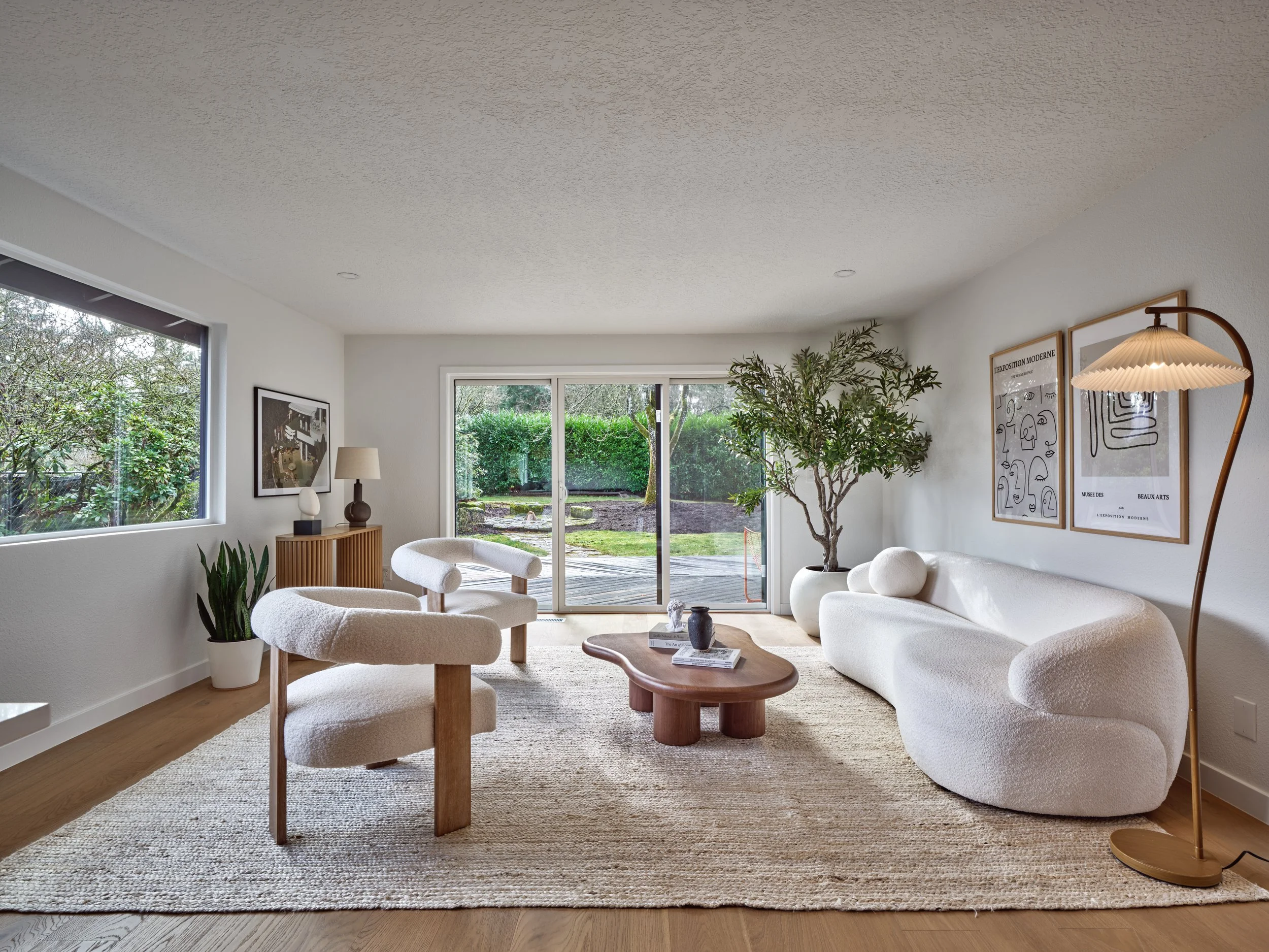 A living room with large windows, a white sofa, two white armchairs, a wooden coffee table with books, a tall potted plant, framed modern art on the walls, a floor lamp, and a view of a lush backyard through a sliding glass door.