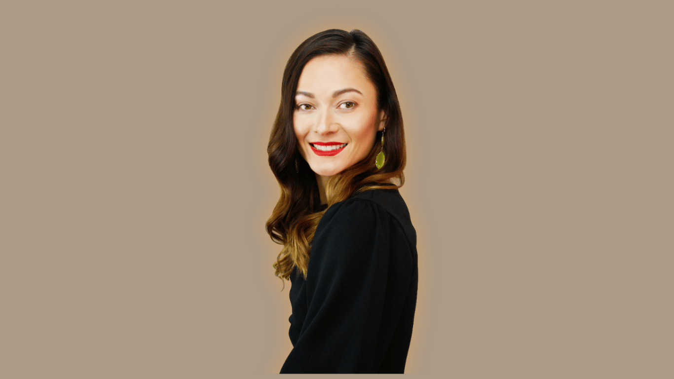 A woman with long wavy brown hair, smiling, wearing a black top and green earrings, standing against a beige background.