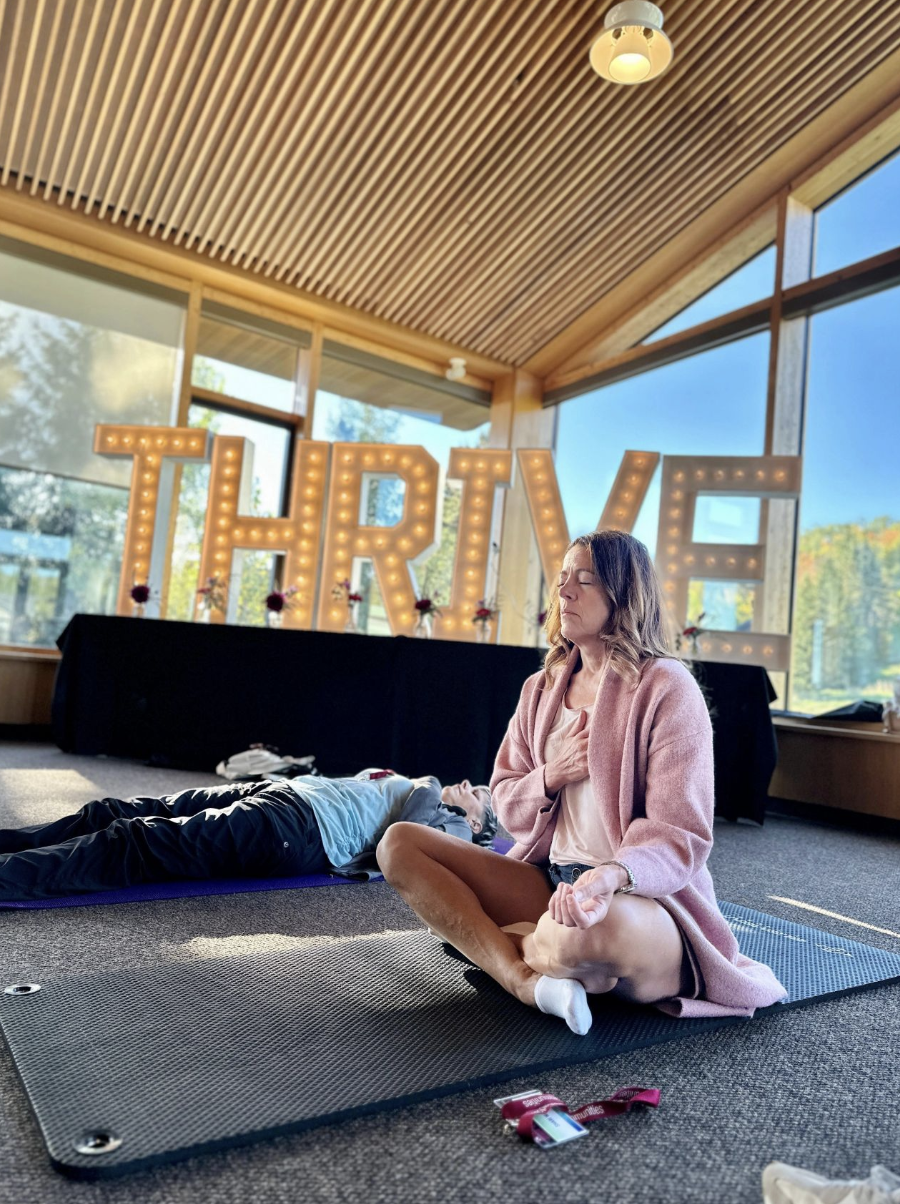 A woman practicing meditation on a yoga mat with her eyes closed and hand on her chest, while a person lies down on another yoga mat nearby. Large illuminated letters spelling 'THRIVE' are in the background, along with a table decorated with small flower arrangements, inside a modern wooden building with large windows and a slanted ceiling.