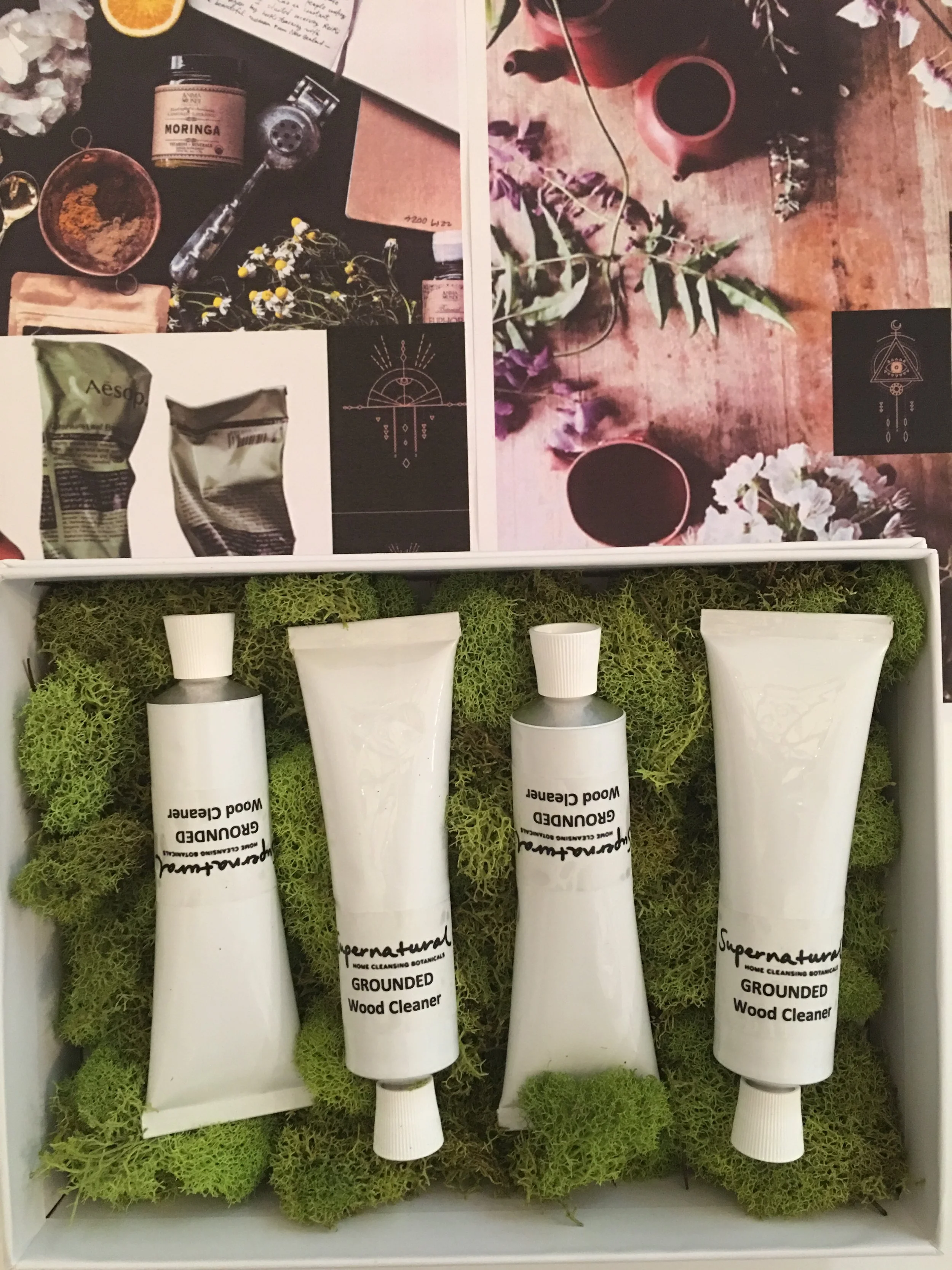 Handmade natural wood cleaner products arranged on green moss in a display box, with paper labels, in front of botanical-themed posters and a wooden table with a teapot and flowers.