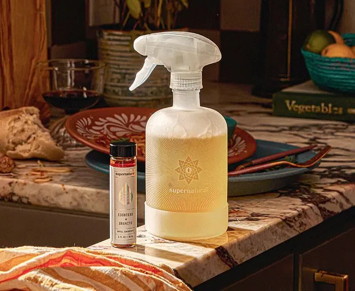 A kitchen countertop with a spray bottle labeled 'supernatural' and a small red container, with a bowl of fruit and other kitchen items in the background.