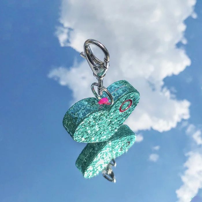 Close-up of a teal glittery airplane-shaped keychain with a metal clasp, hanging against a partly cloudy blue sky.