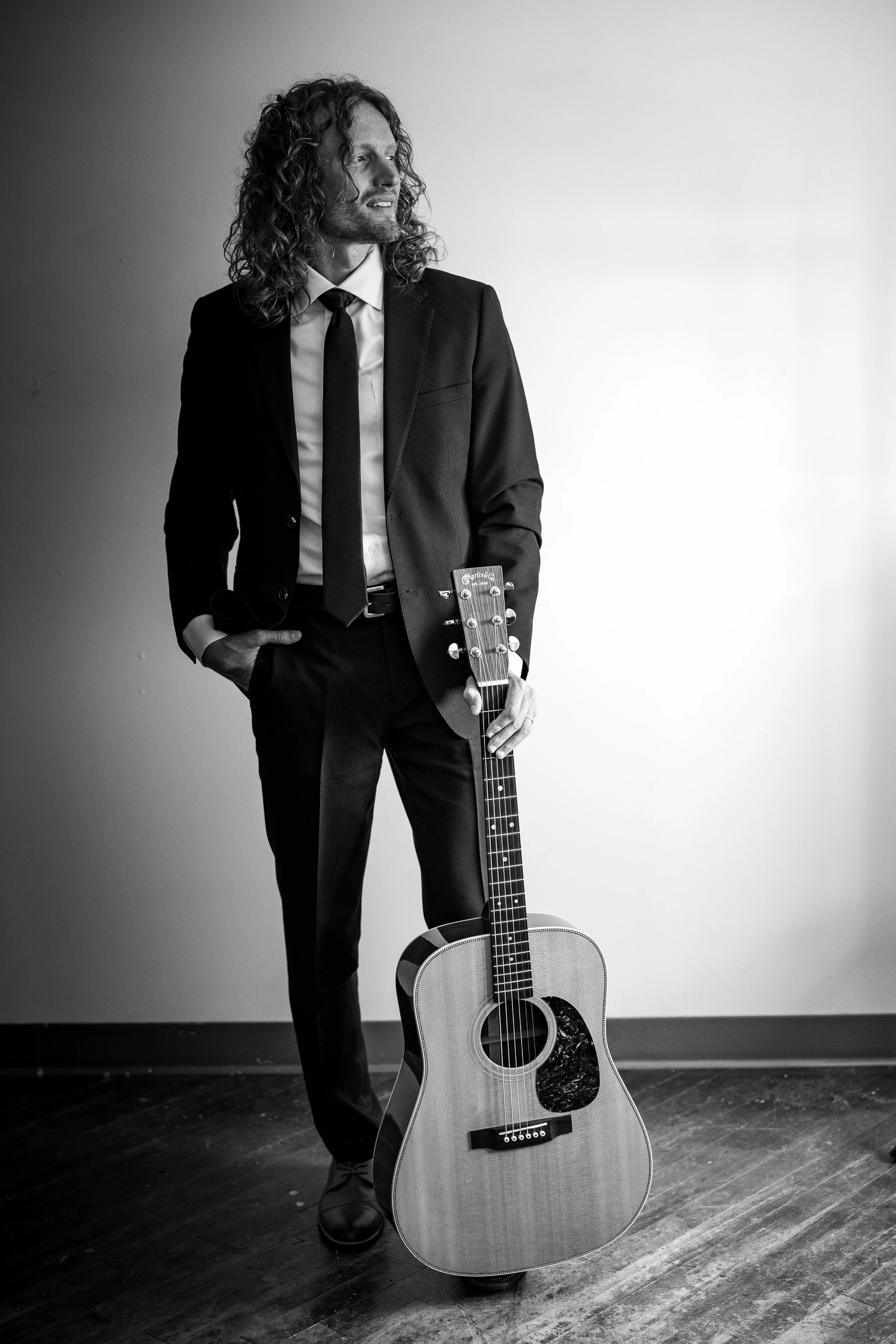 Christopher Burkholder in a suit holding his acoustic guitar, standing on a wooden floor against a plain white wall. The photo is in black and white.