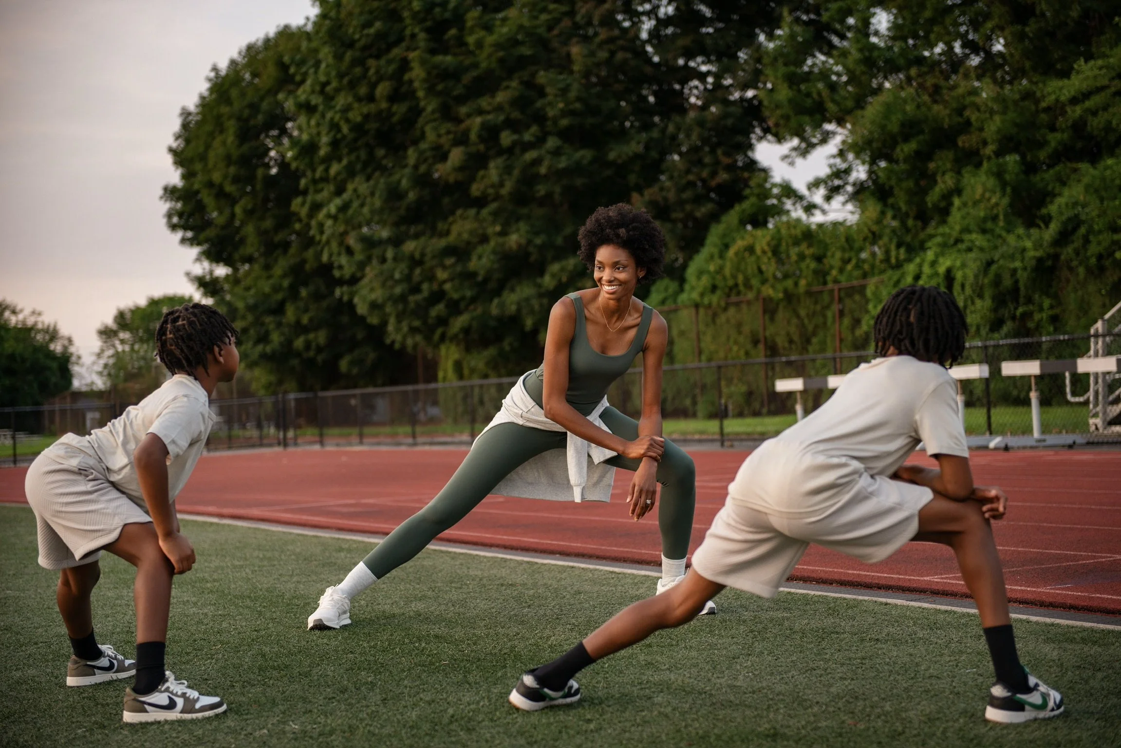 SoulShape founder leading exercise with her two children on a sports field with a track and green trees in the background during the evening.