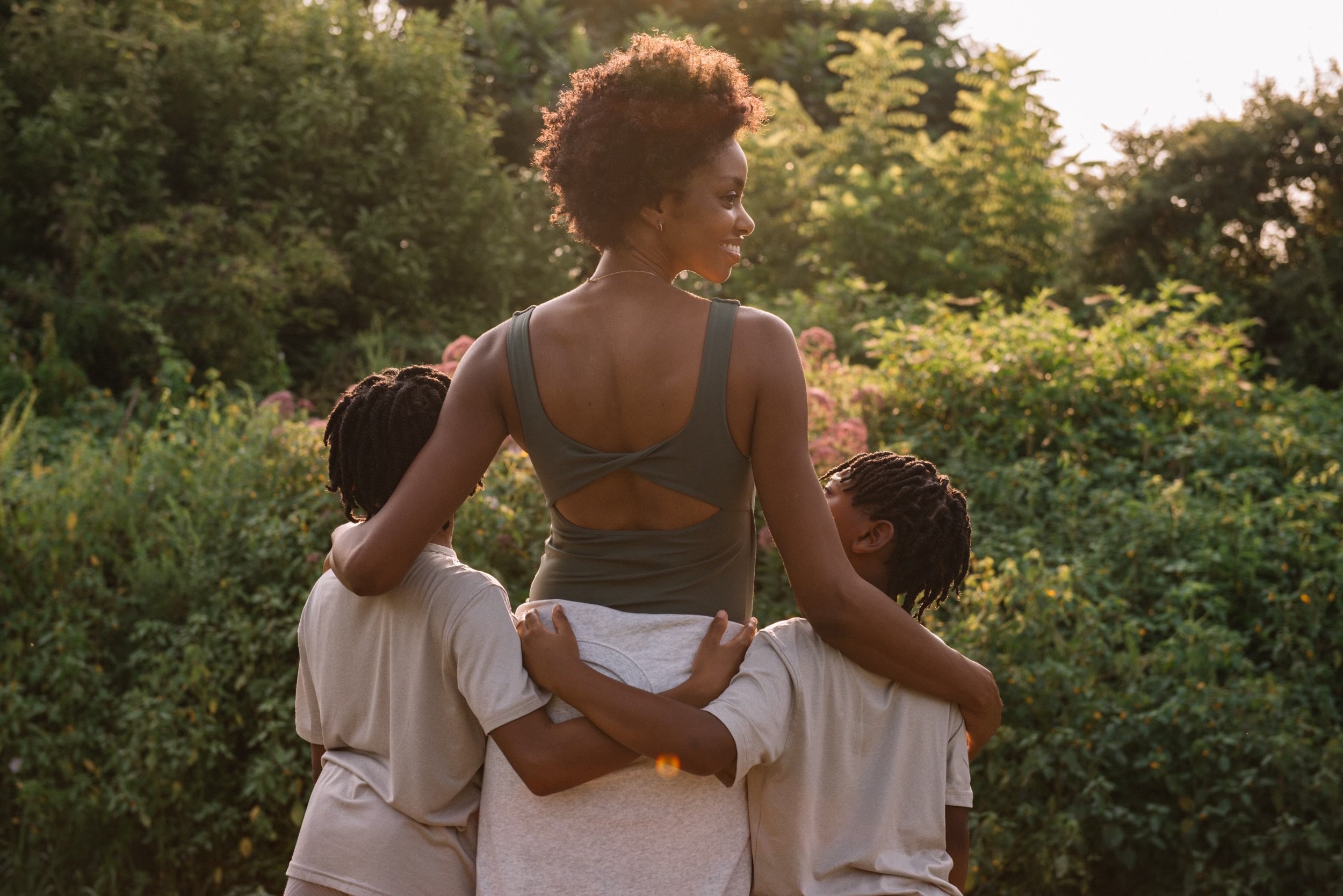 Black mother in a green sports bra walking outdoors, embracing her two sons, in a lush garden at sunset.