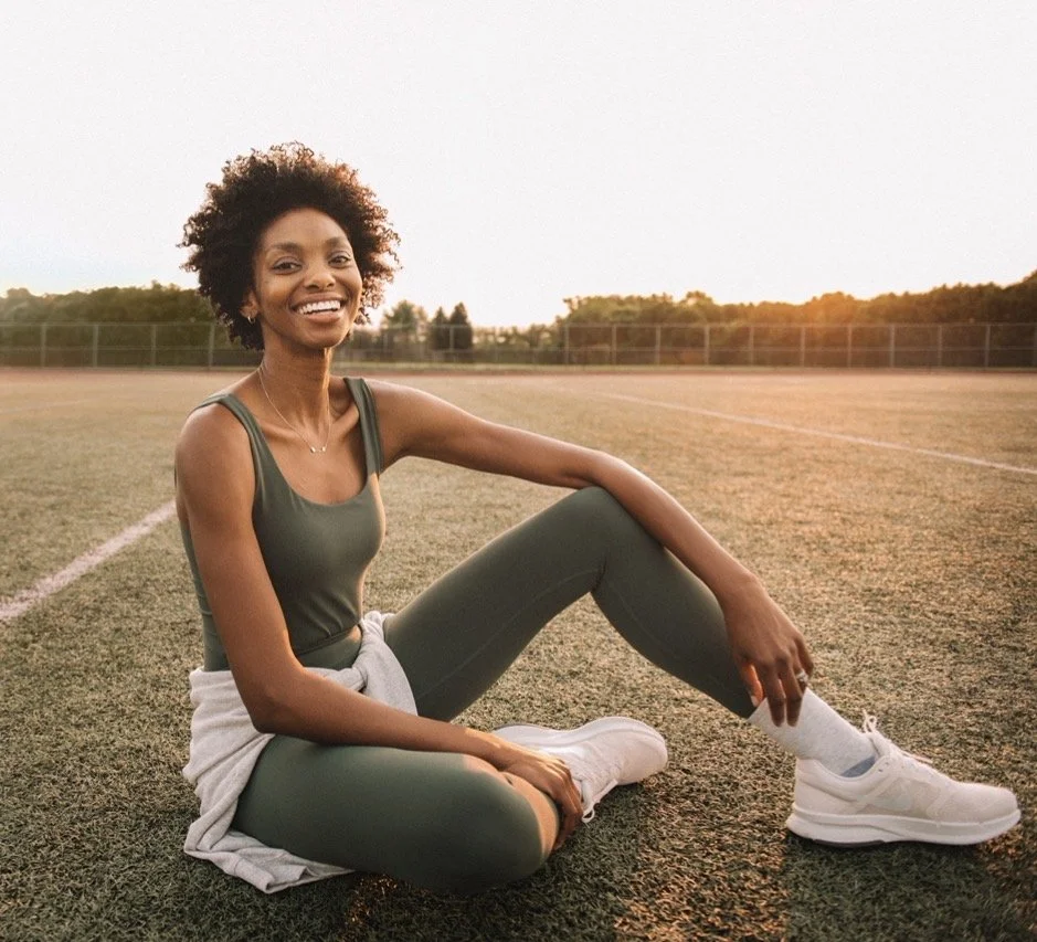 A woman with natural hair, wearing a green athletic outfit and white sneakers, sitting on a sports field during sunset, smiling at the camera.