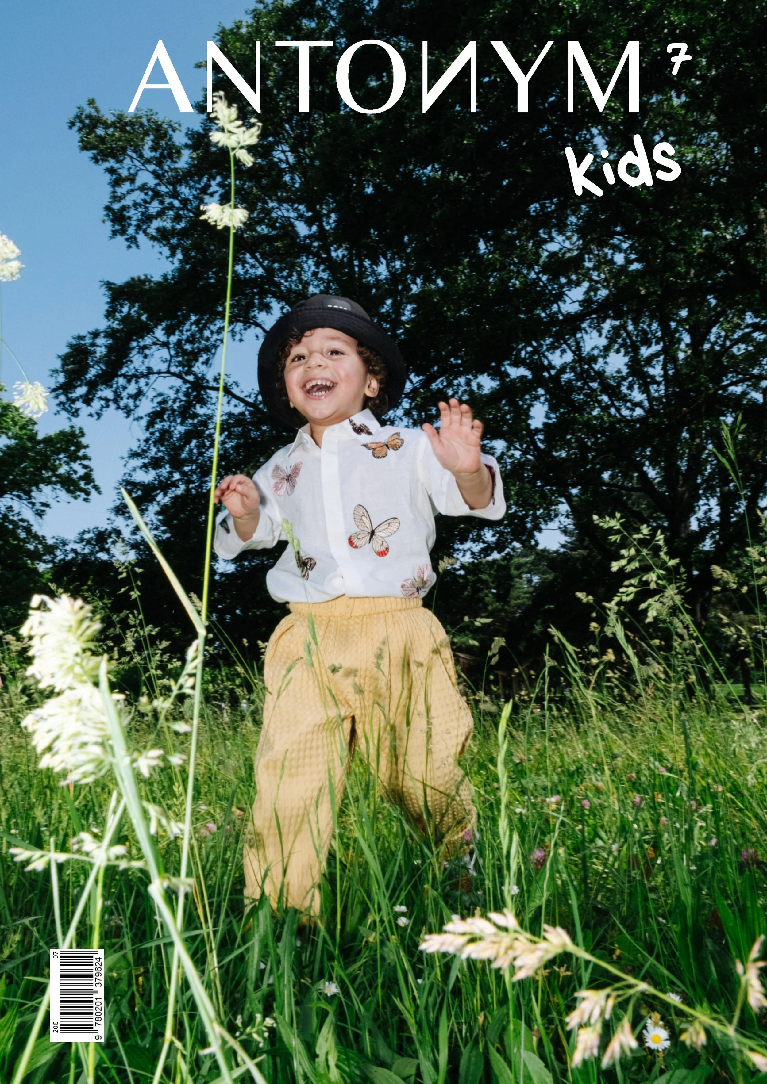 Jeune enfant souriant, portant un chapeau noir et une chemise blanche à imprimé de papillons, debout dans un champ d'herbe et de fleurs, avec un grand arbre en arrière-plan, sous un ciel bleu.