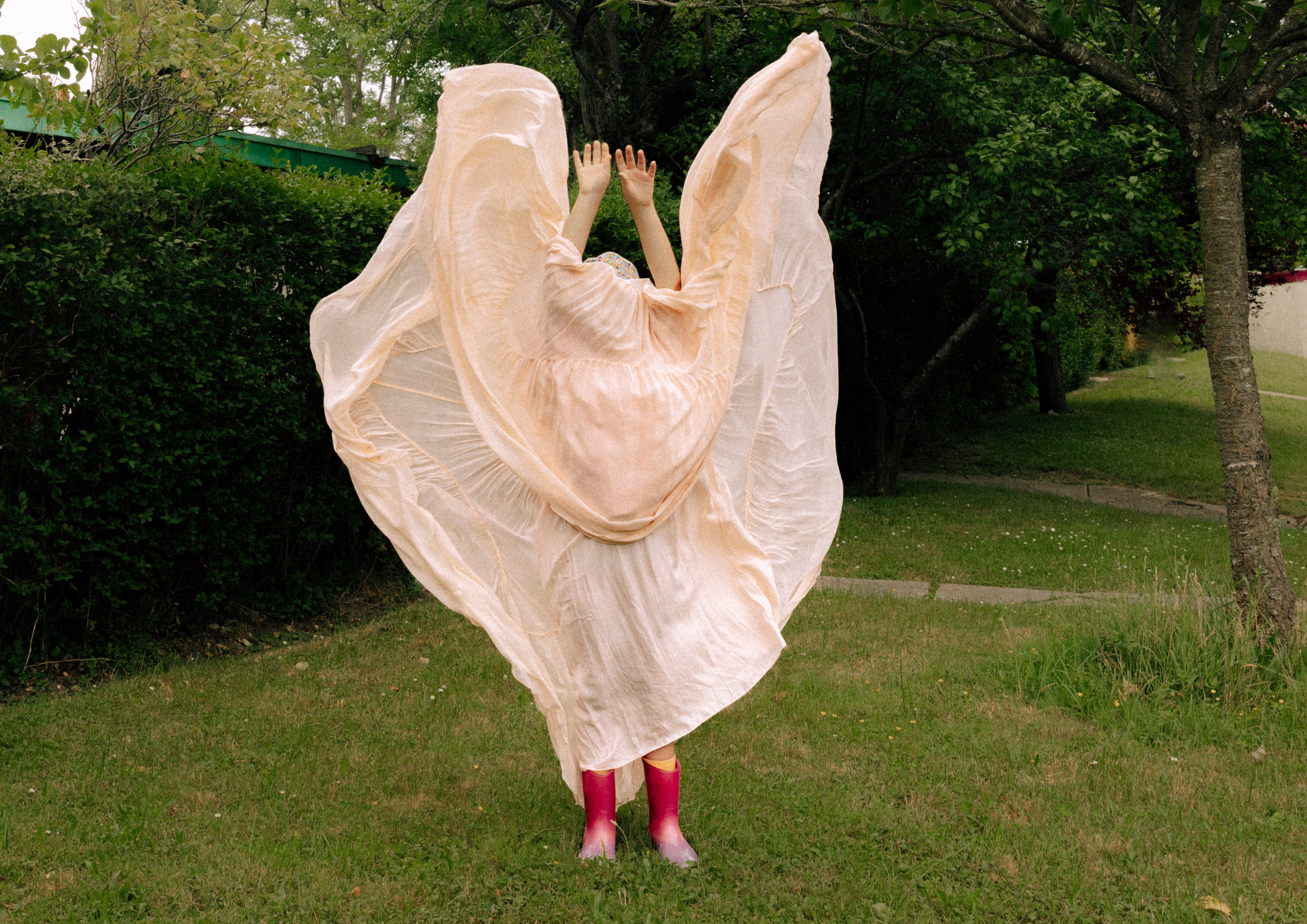 Une fillette vêtue d'une robe fluide de couleur beige, avec de grands plis, porte des bottes rose vif et se tient dehors dans un espace verdoyant avec des arbres et un ciel partiellement visible.