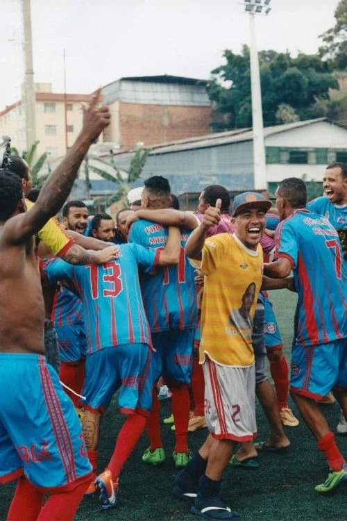 Soccer players celebrating together on the field, with one player smiling and wearing a yellow jersey with the number 2, and others in blue and red uniforms.