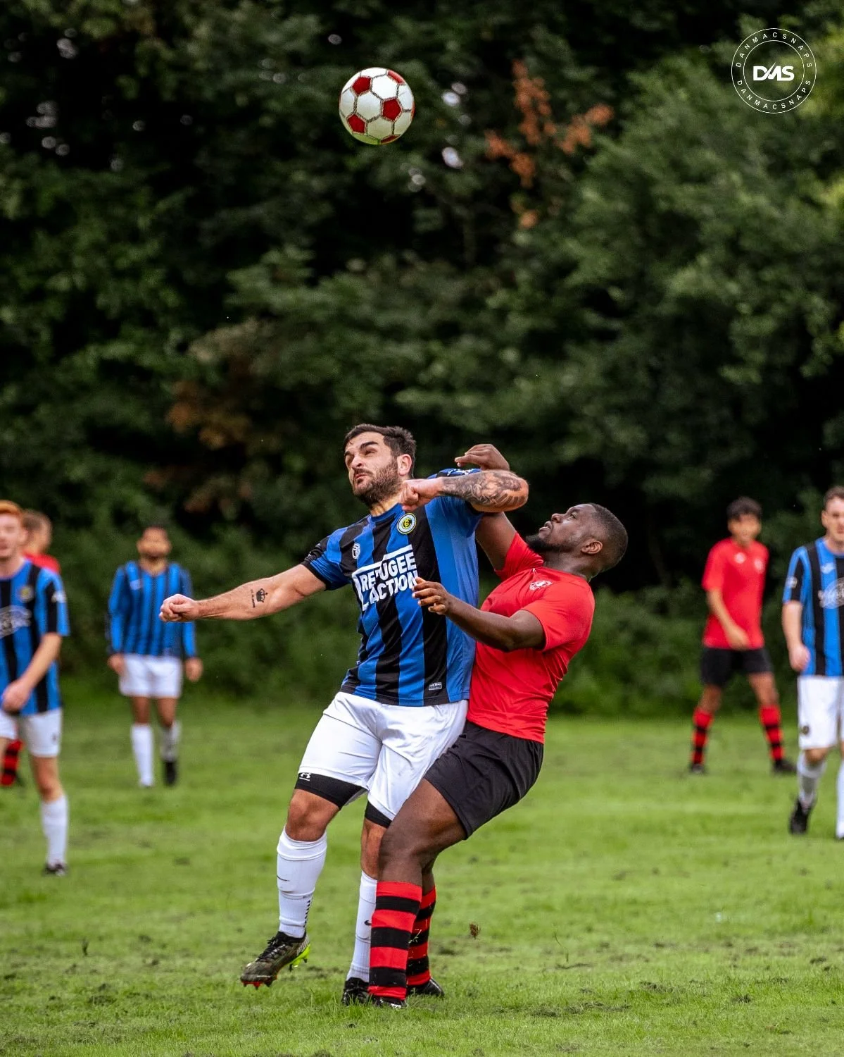 Two soccer players are jumping for the ball during a game, with one in a blue and black striped jersey and the other in a red jersey. Several other players are visible in the background on a grassy field with trees.