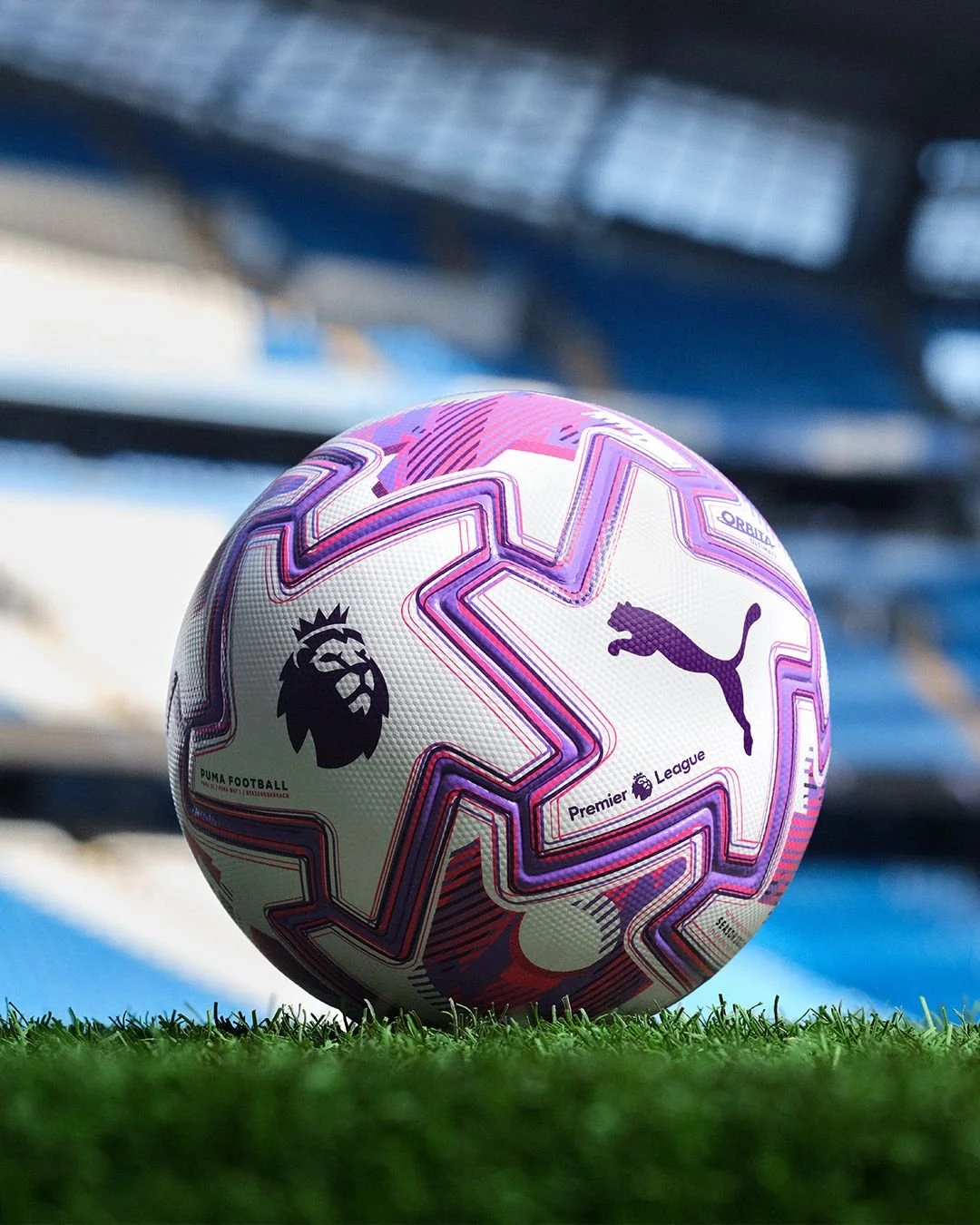 A white soccer ball with pink, purple, and black star patterns, featuring the Premier League logo, Puma logo, and a lion logo, resting on green grass with a blurred stadium background.