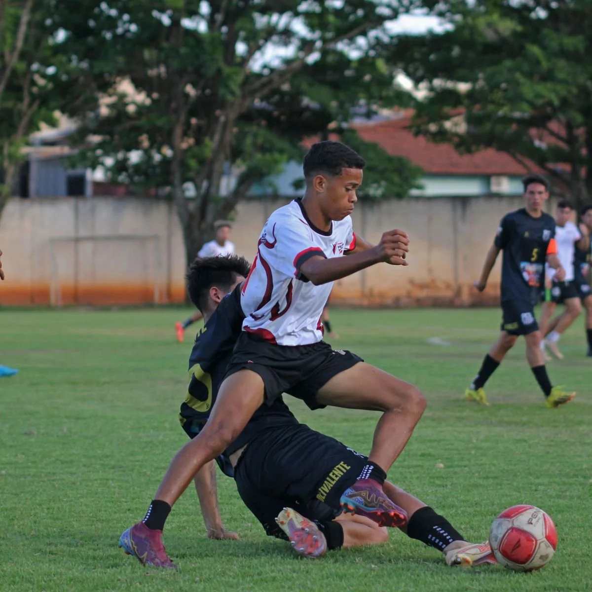 Two young soccer players competing for possession of the ball during a match, with other players and trees in the background.