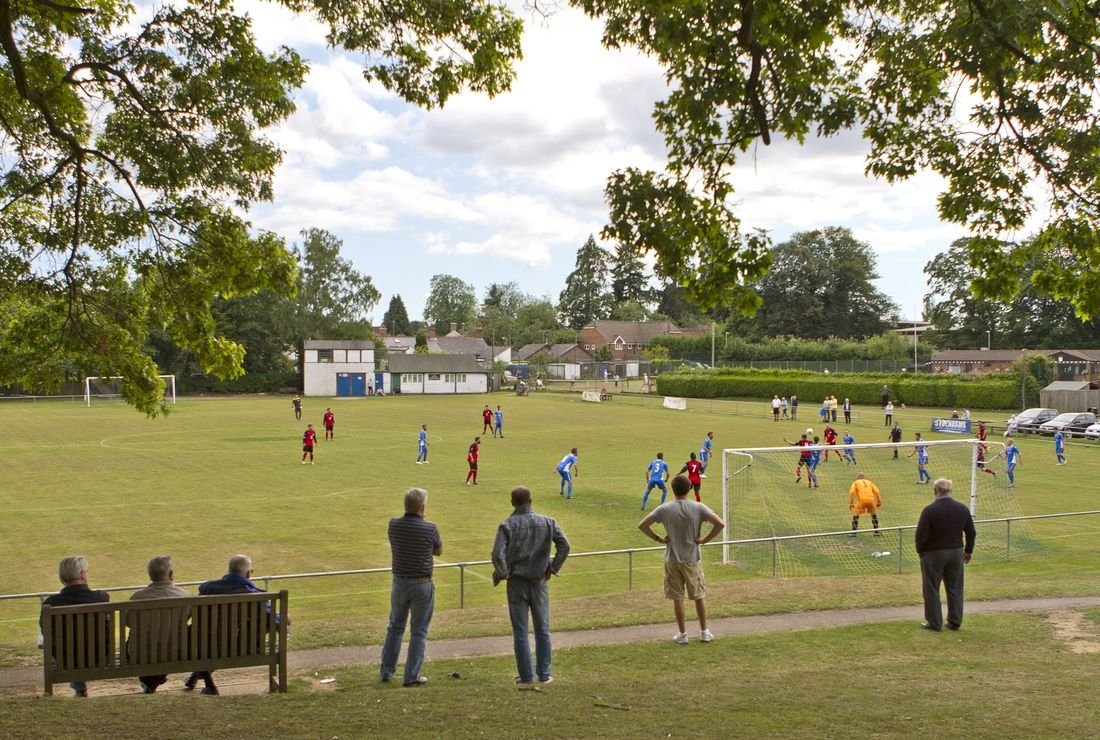 People watching a soccer match on a grassy field from a fenced area with benches, trees, and houses in the background.