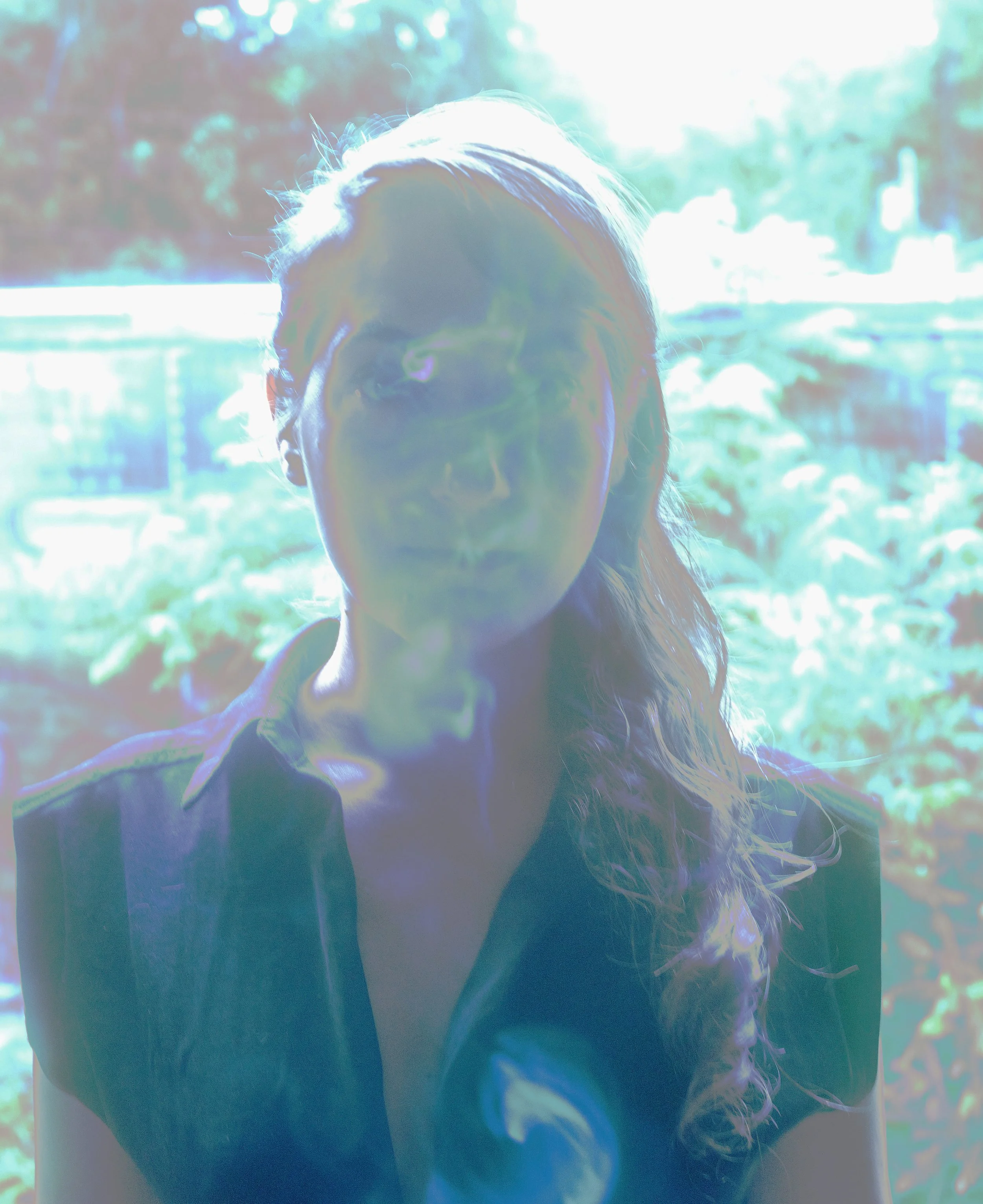 A person with long hair standing outdoors with a bright backlit background and some trees visible, their face is partially shadowed.