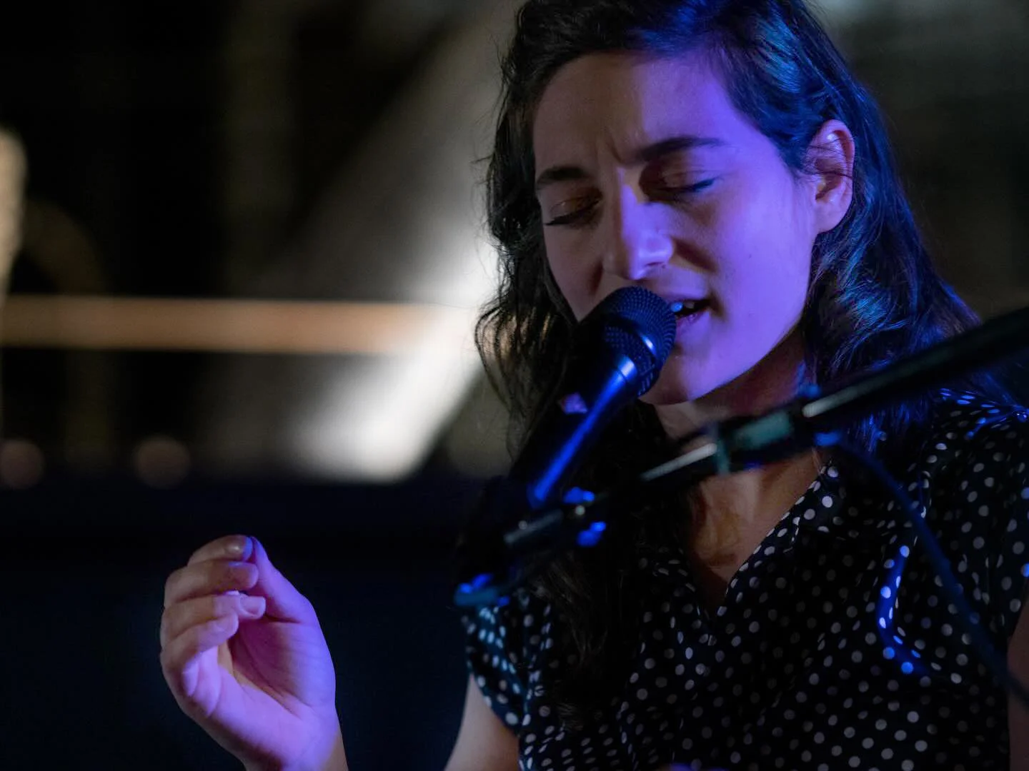 A woman with dark, wavy hair sings into a microphone while playing a piano, wearing a black polka dot top, in a dimly lit setting.