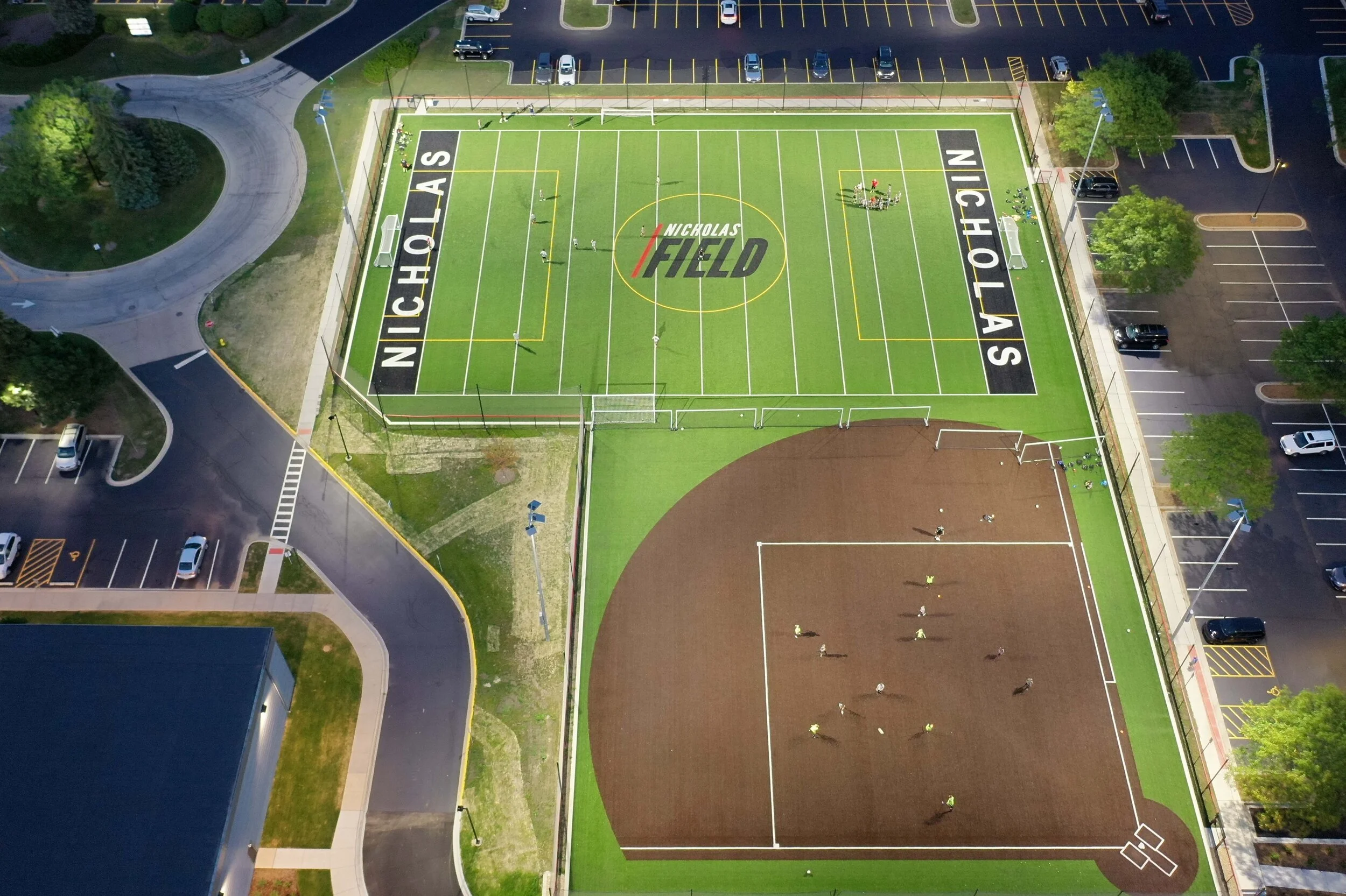 Aerial view of Nicholas Field, a multi-use sports field with a green grass surface, marked for soccer, with parking lots and surrounding trees and pathways.