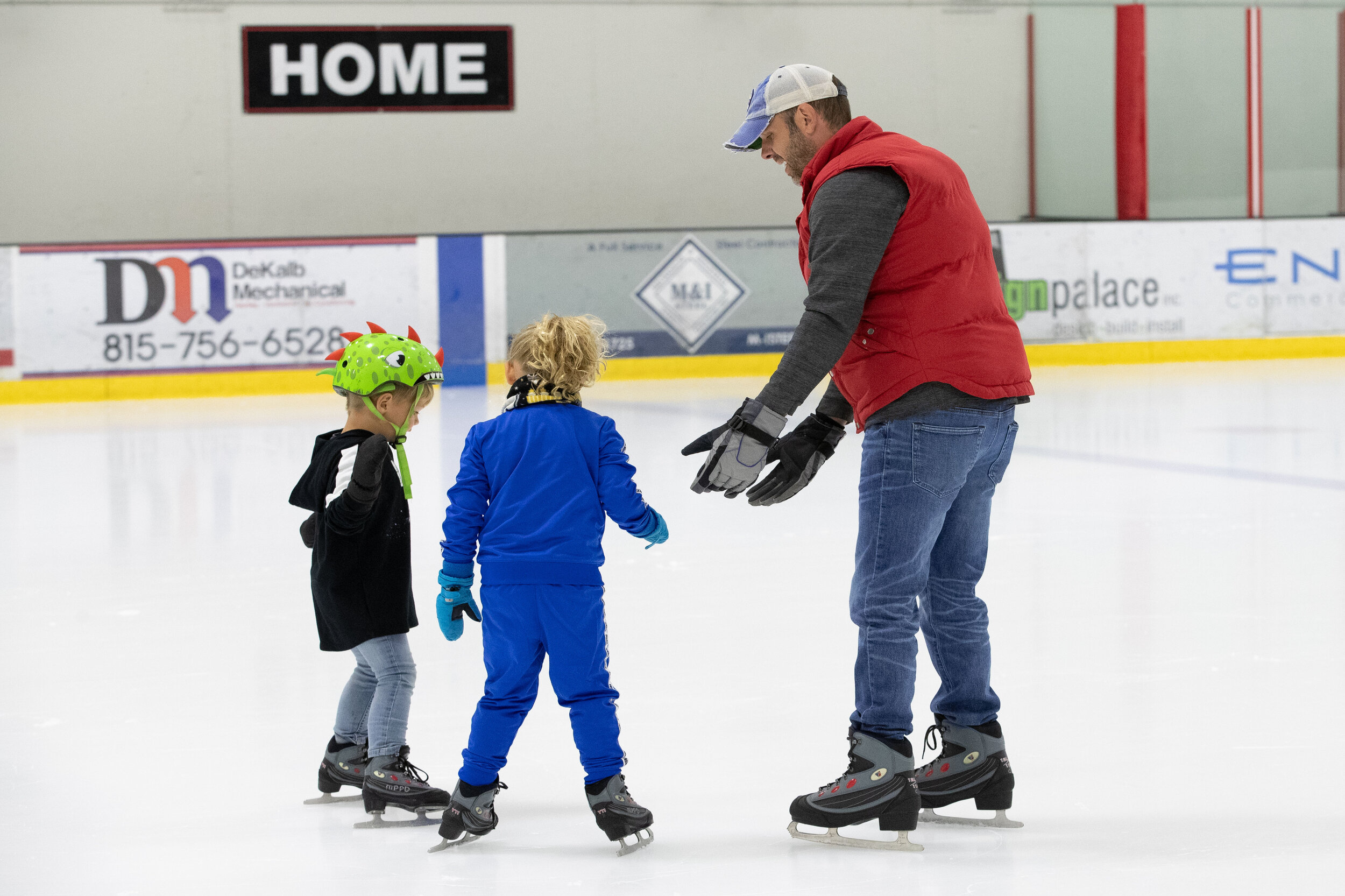 An adult man teaching two young children how to ice skate at an indoor ice rink. The children are wearing helmets and ice skates, and the man is guiding them.