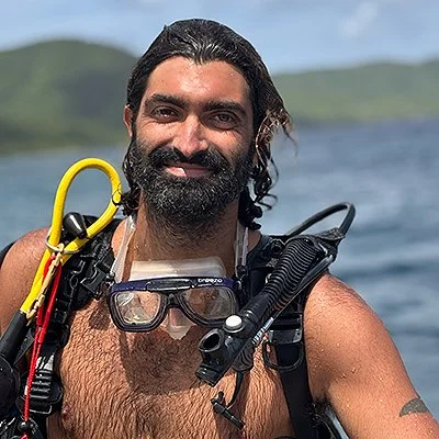 Man with black hair and beard smiling outdoors with a body of water in the background, wearing scuba gear including a mask, snorkel, and wetsuit.
