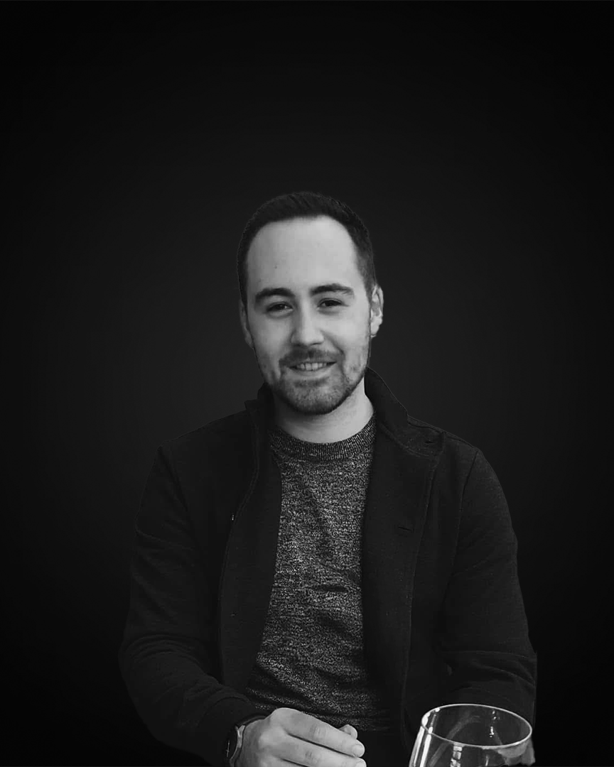A black and white portrait of a young man sitting at a table, smiling, with a glass in front of him against a solid dark background.