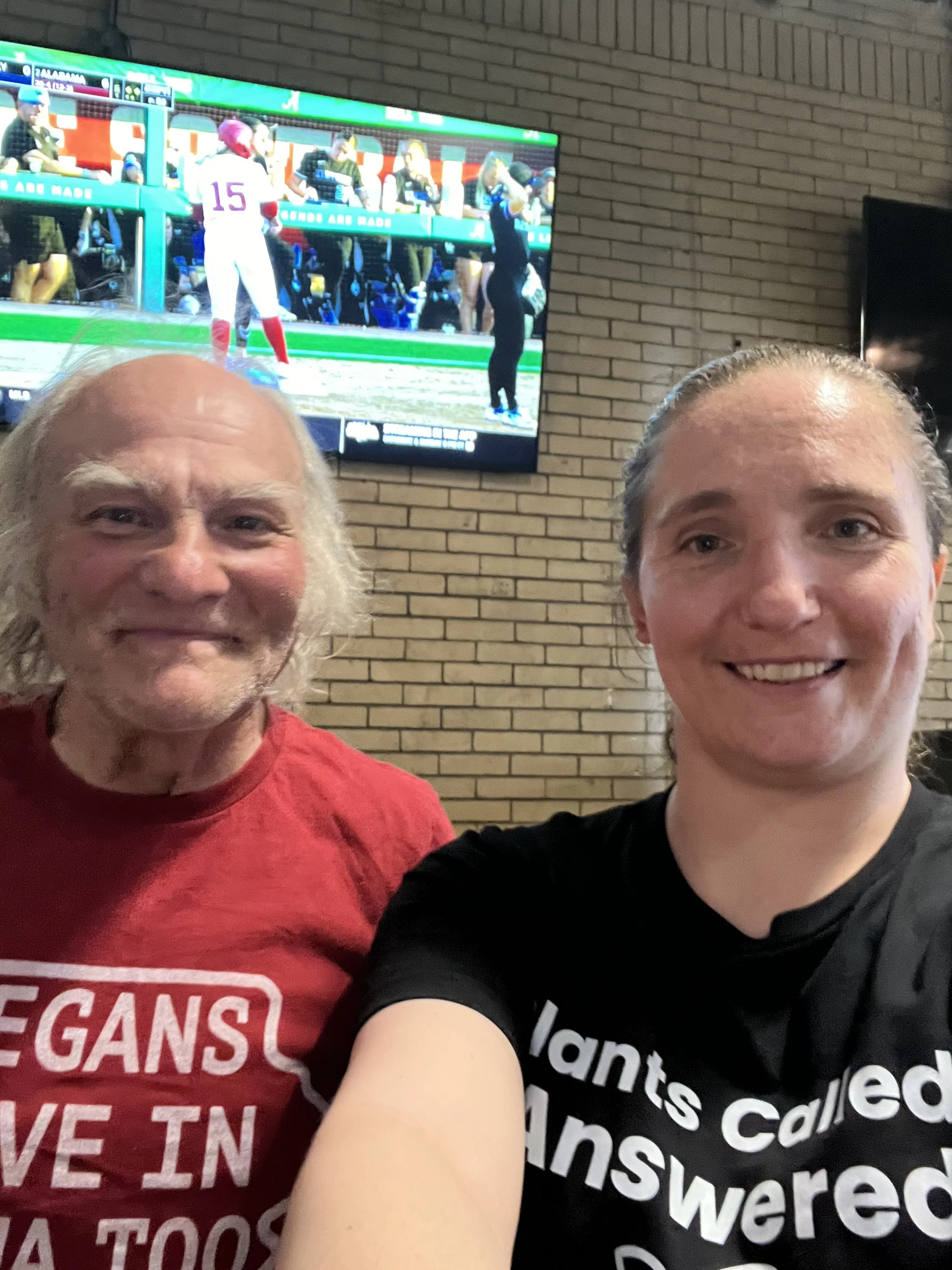 Two smiling women taking a selfie in a restaurant with a brick wall and a television screen showing a football game in the background.