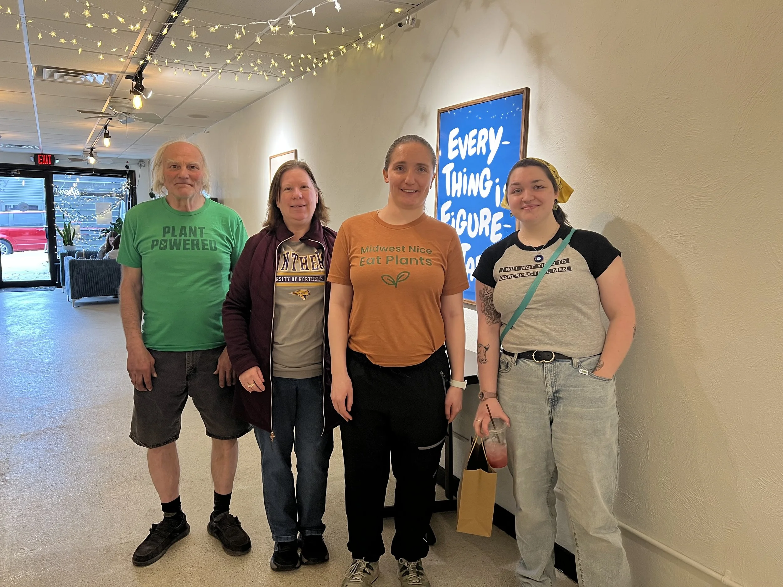 Four people standing indoors in front of a wall with lights and a sign that says "Every-Thing is Figure",