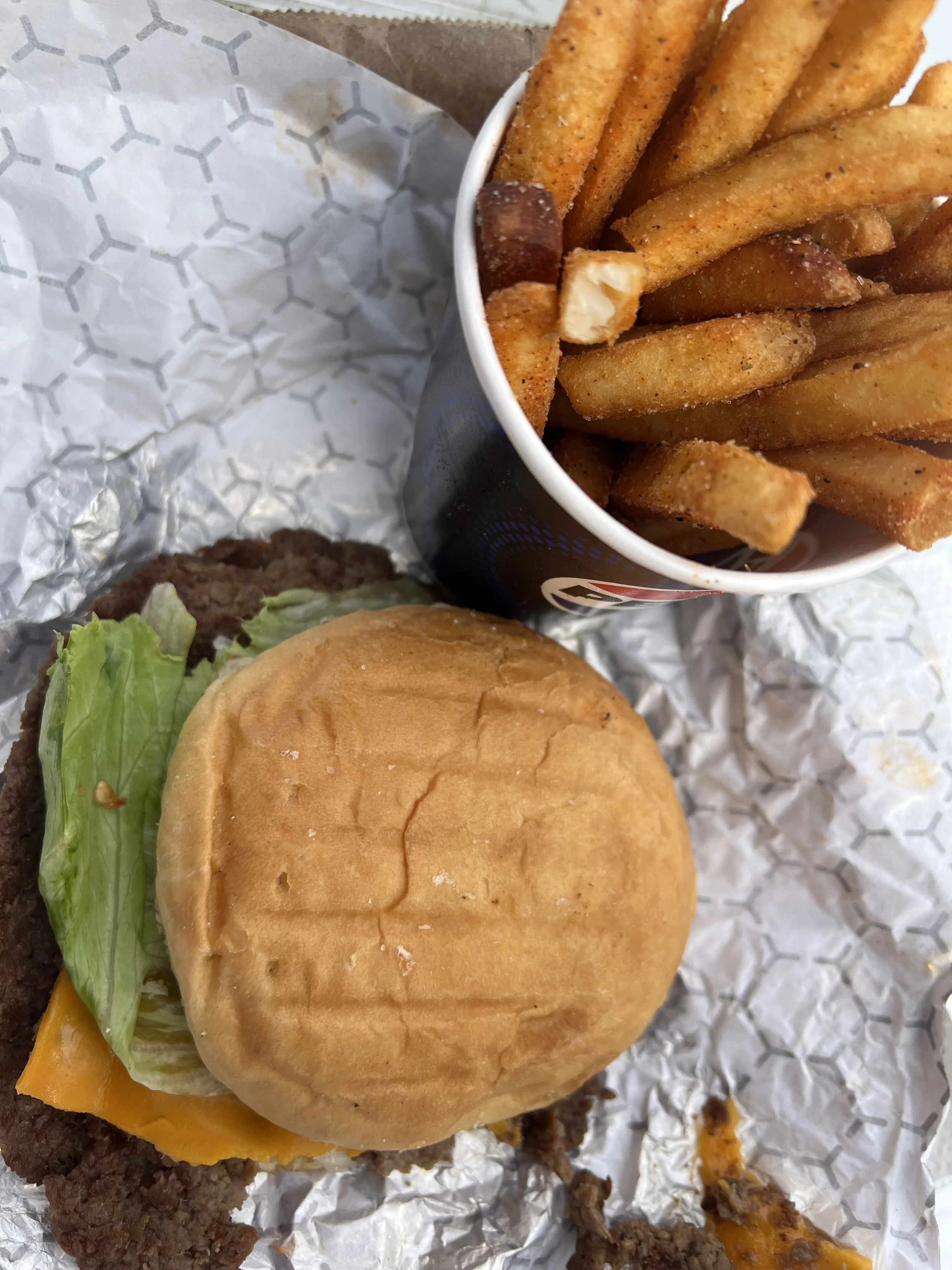 Close-up of a burger with lettuce, cheese, and a bun, alongside a cup of French fries.