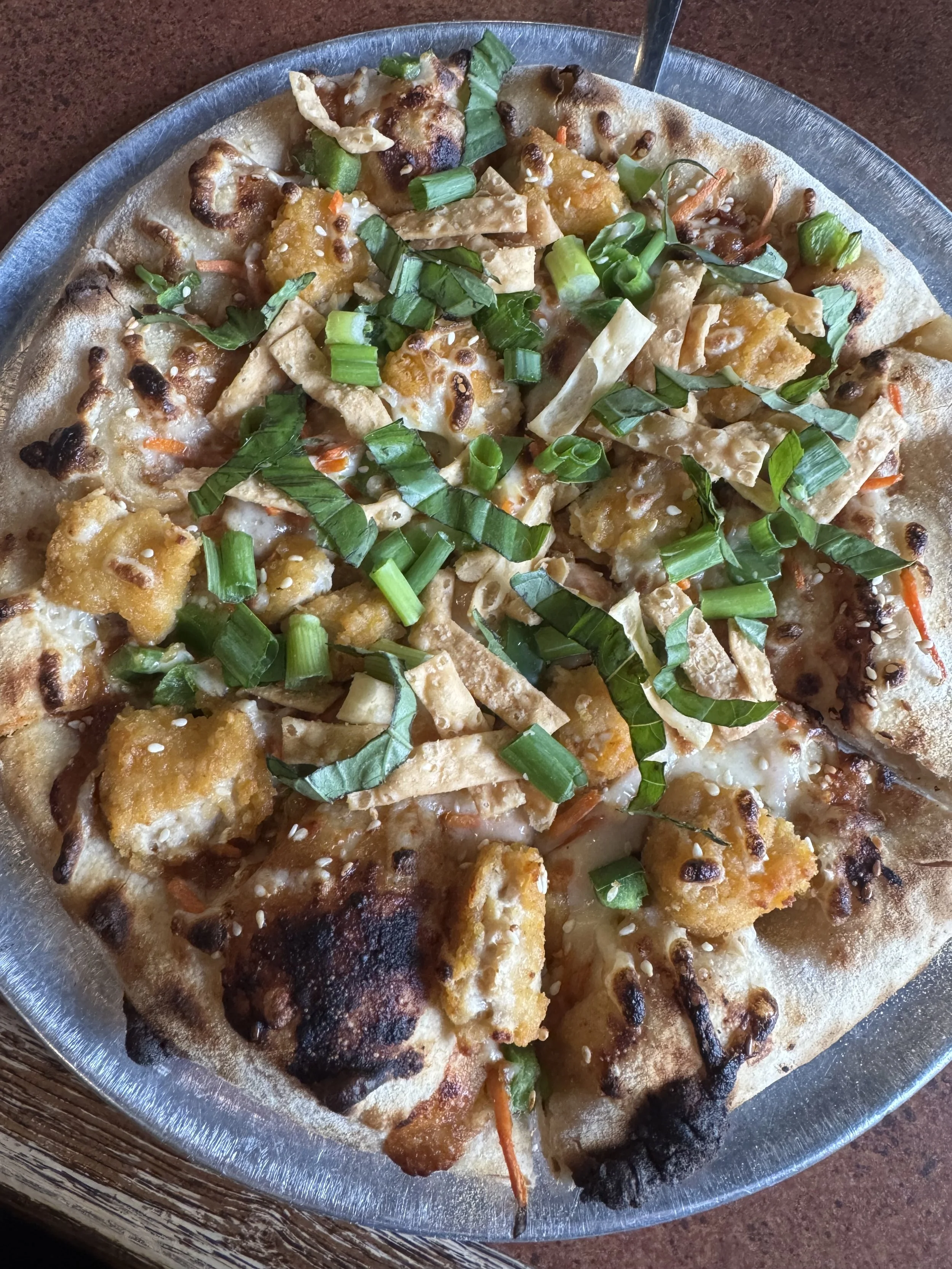 A close-up view of a pizza with toppings including chopped green onions, sesame seeds, fried chicken pieces, and vegetables on a metal plate.