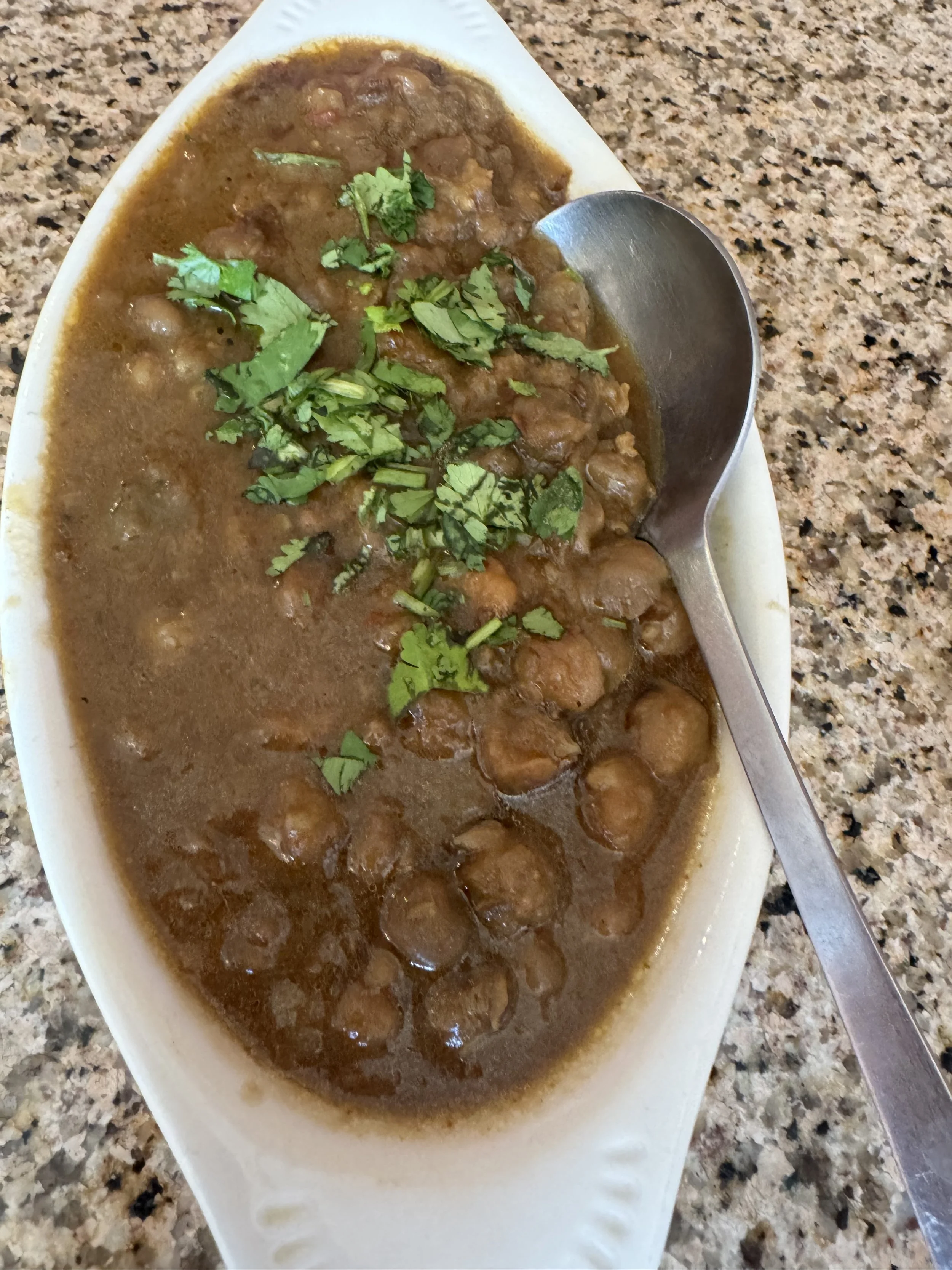 A white dish of brown stew garnished with chopped cilantro on a granite countertop with a metal spoon resting inside.