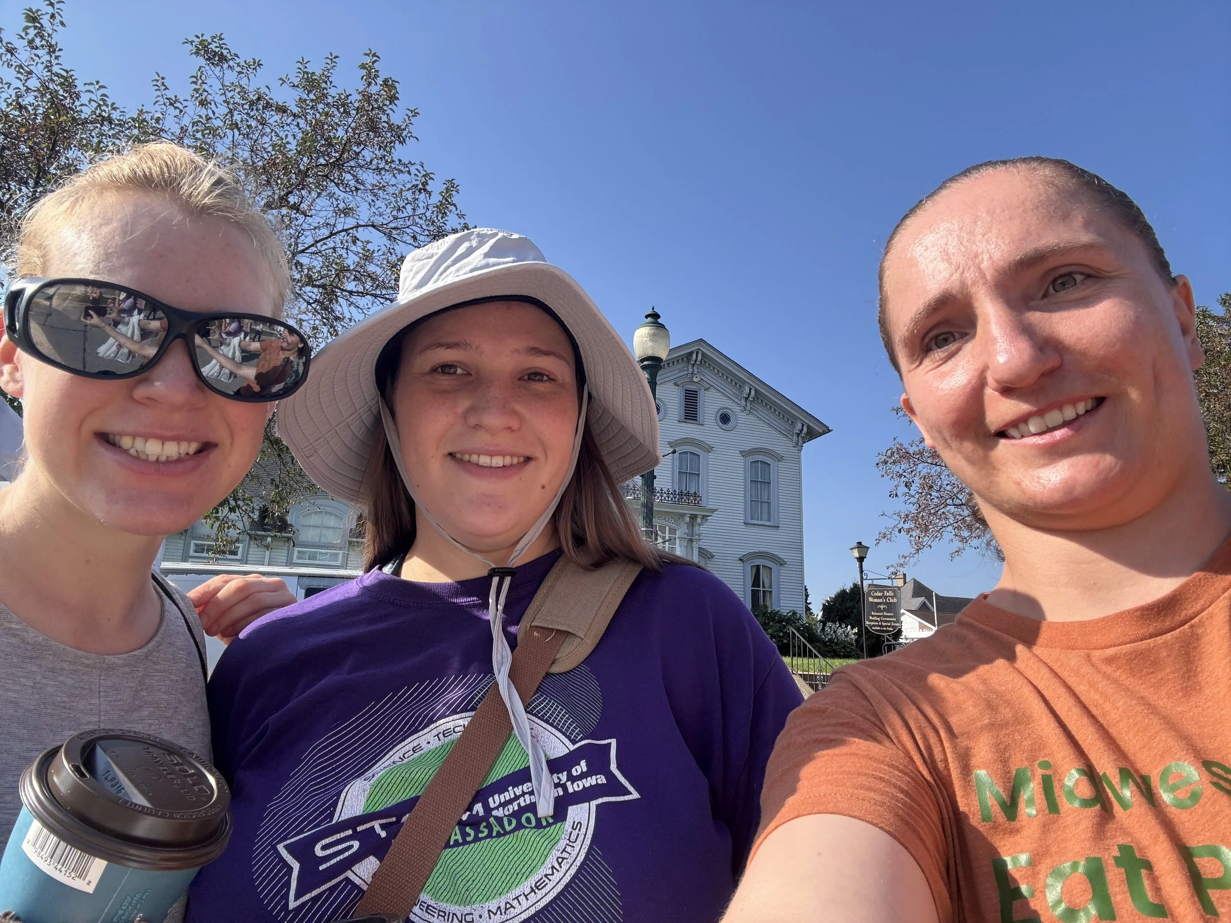 Three young women taking a selfie outdoors on a sunny day, with a blue sky, trees, and a historic white wooden building in the background.