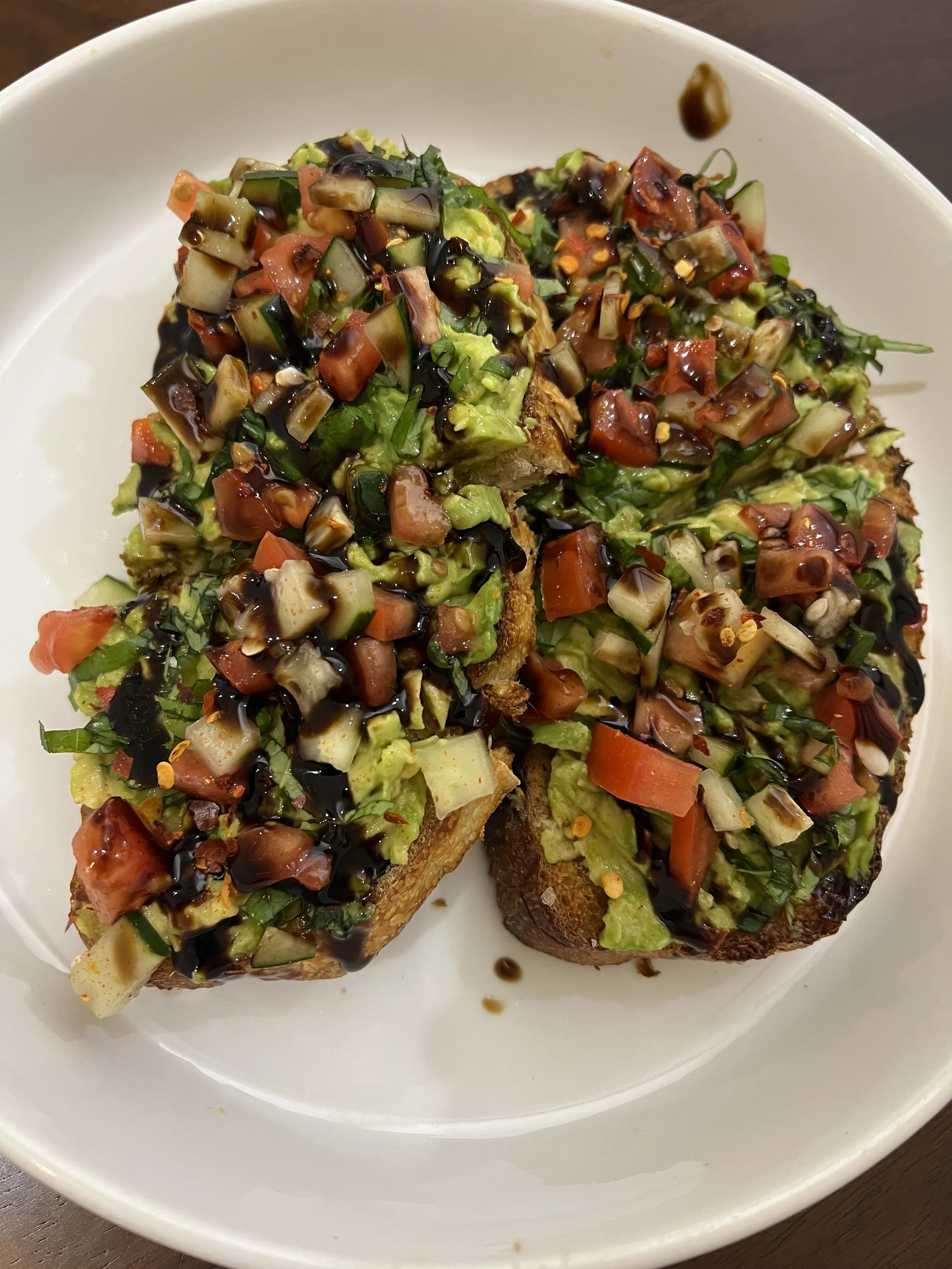 Close-up of toasted bread topped with chopped tomatoes, cucumbers, mixed greens, and drizzled with balsamic glaze, served on a white plate.