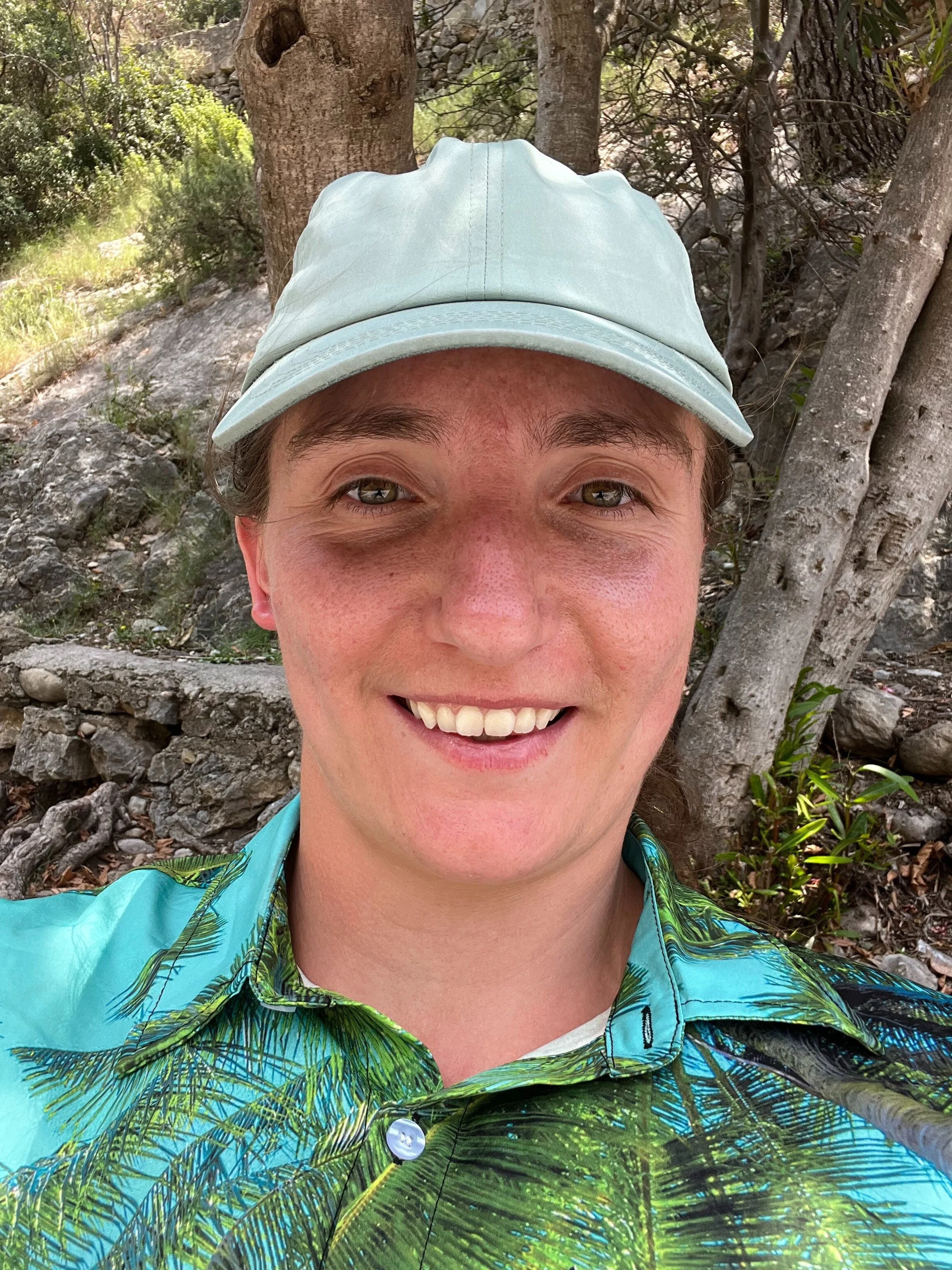 A person outdoors wearing a light blue cap and a tropical patterned shirt, with trees and rocks in the background.