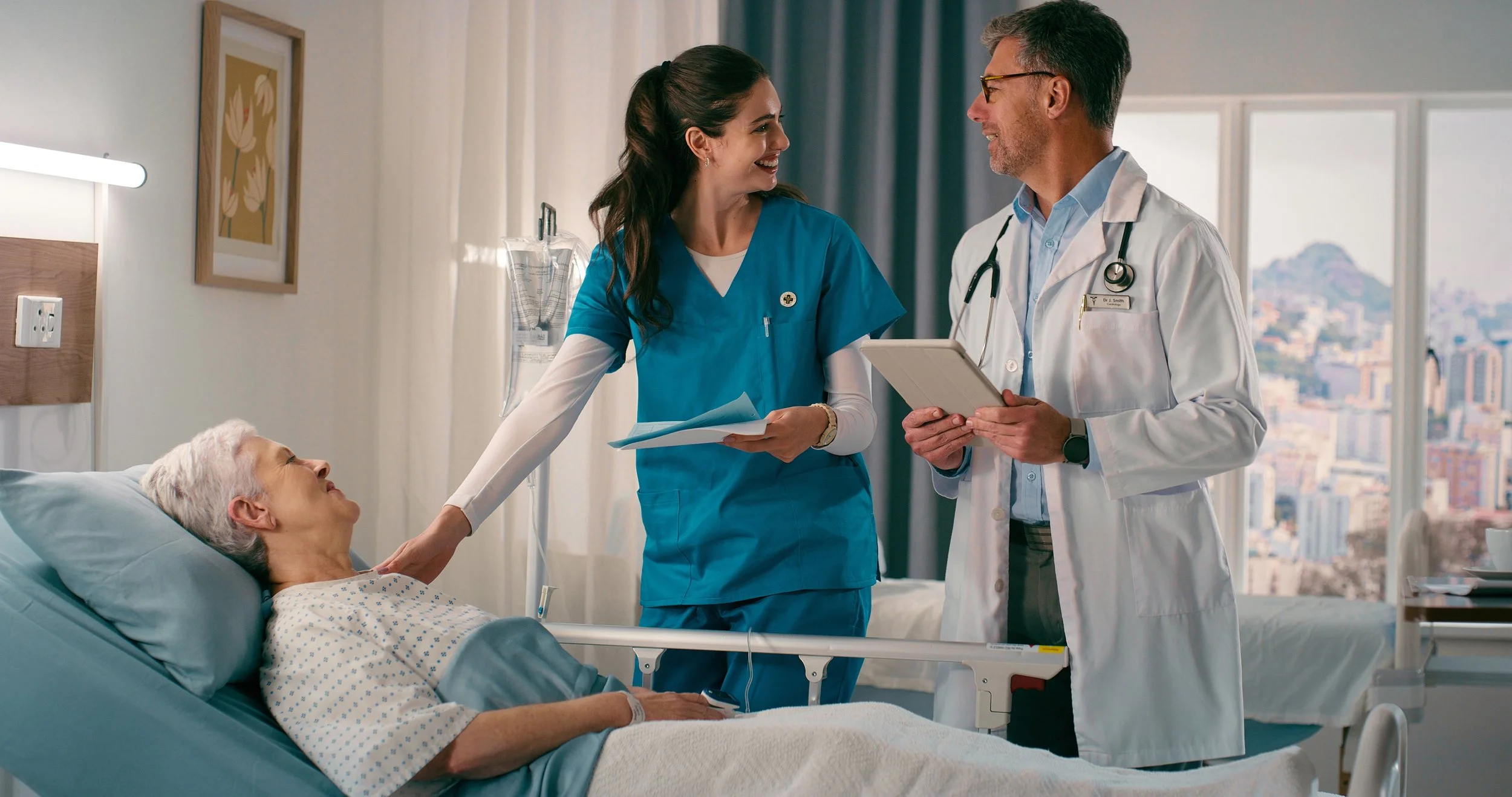 An elderly woman is lying in a hospital bed, talking to a female nurse and a male doctor who are standing next to her. The nurse is smiling and holding a clipboard, while the doctor holds a tablet. The room has a large window with a cityscape view.