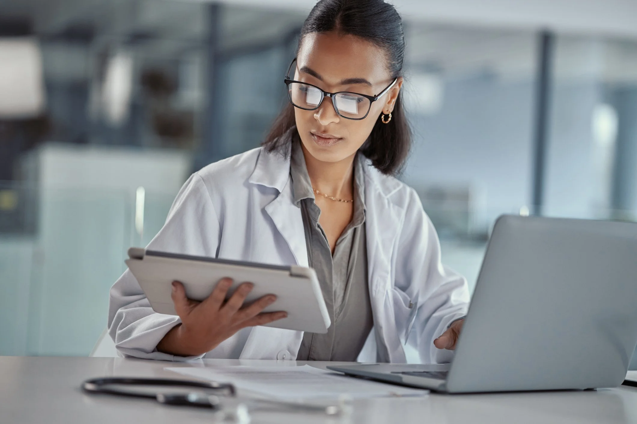 A young woman wearing glasses and a white lab coat is sitting at a table, holding a clipboard and looking at her tablet. She appears to be in a modern office or laboratory setting.