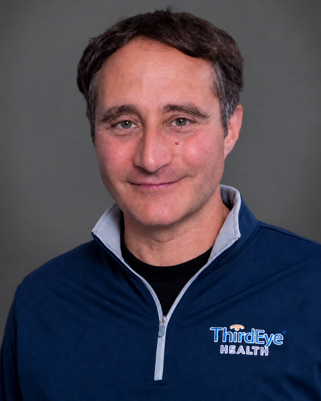 A man with short dark hair, wearing a navy blue jacket with a white zipper and a logo that says "Third Eye Health" on it, smiling against a gray background.